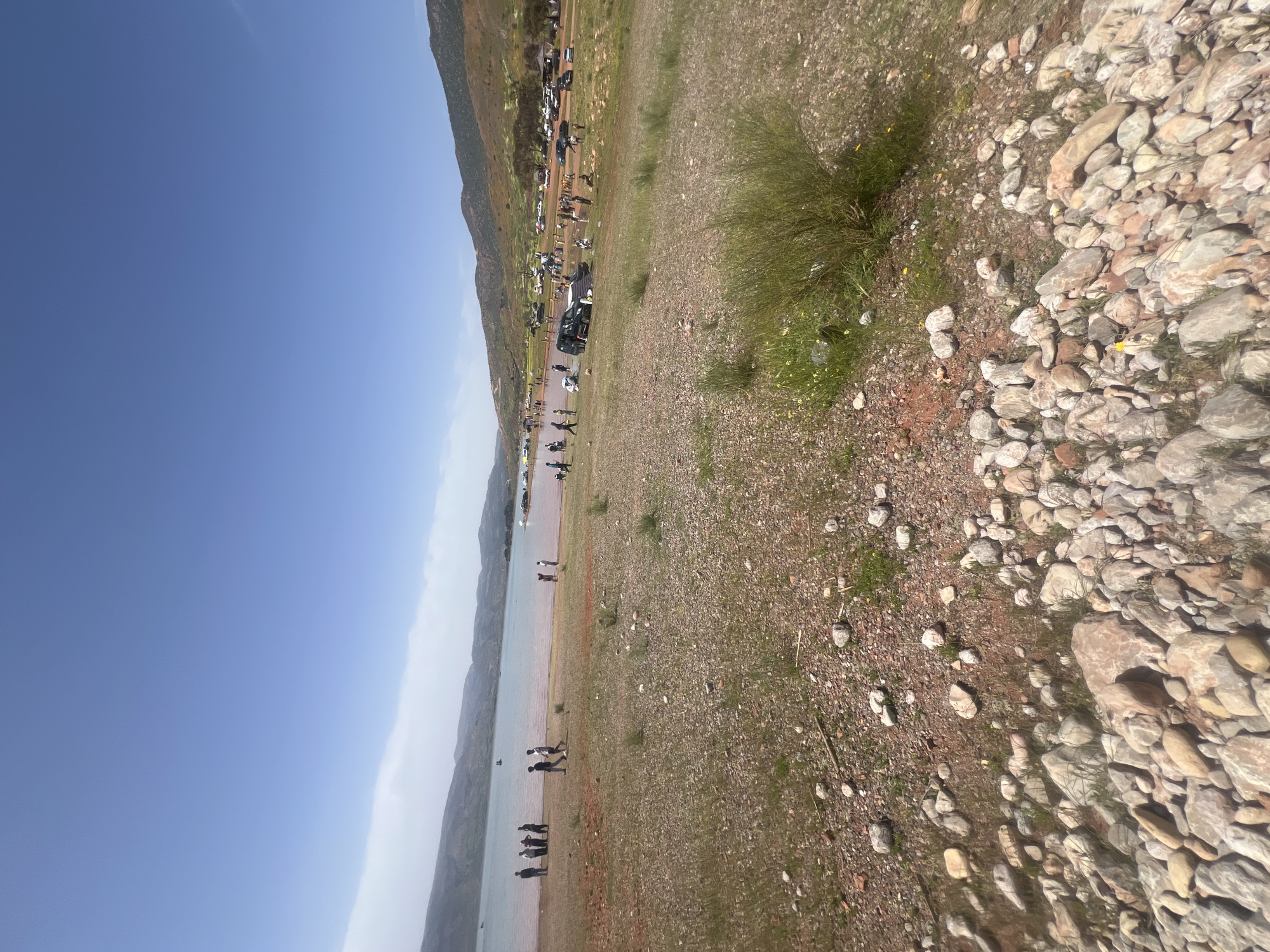 The image depicts a scene at Bin El Ouidane, Morocco, featuring a rocky shoreline with a lake or reservoir visible. People are gathered near the water's edge. Beyond the shoreline, a green, grassy area extends upward towards a line of vehicles parked along the slope of a mountain. The sky is clear and blue. The foreground consists of a mix of rocks and sparse vegetation.