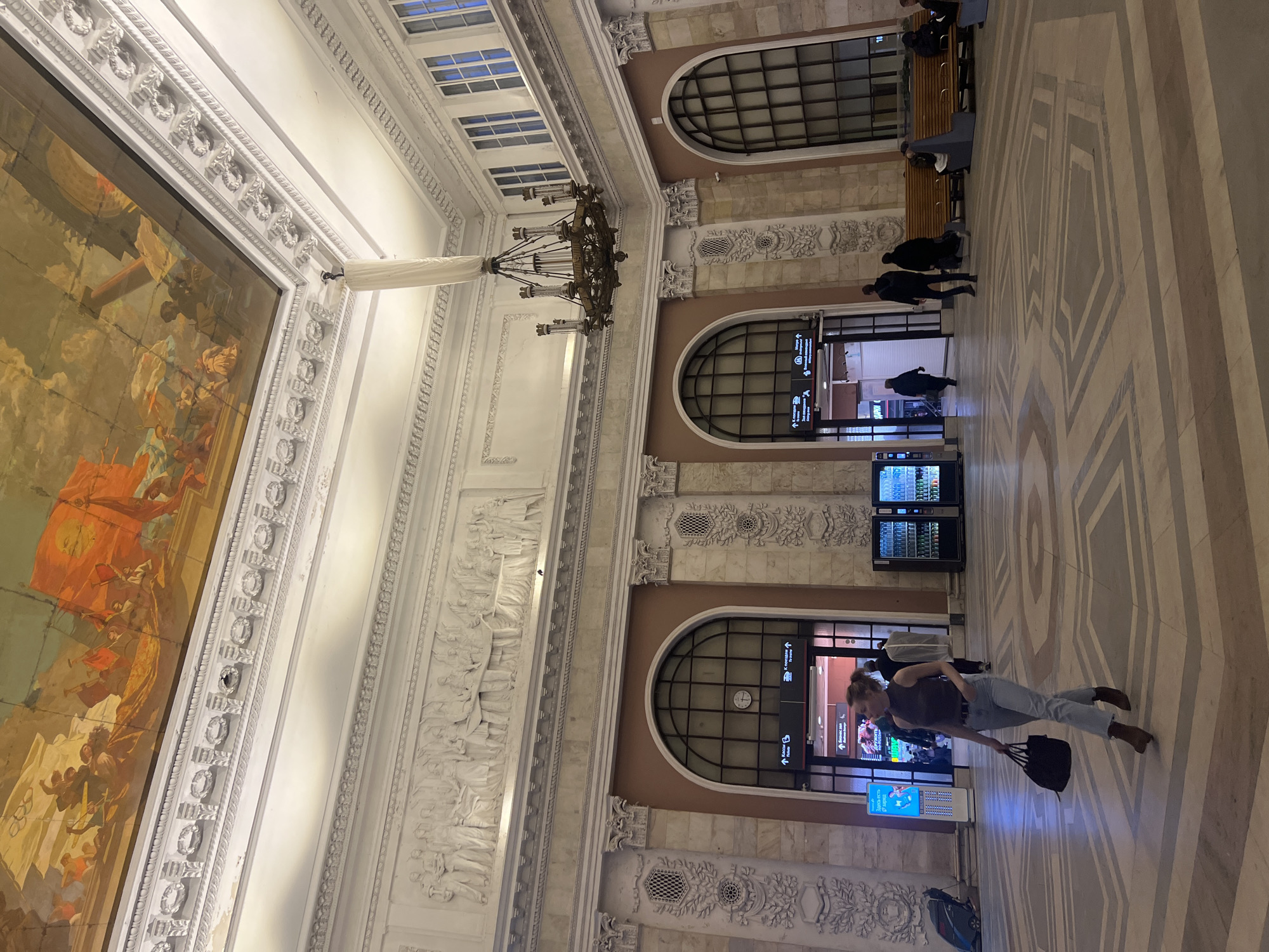 The image captures the interior of a grand building, potentially a railway station, characterized by its high ceilings, ornate architectural details, and marble floors. The location is Saint Petersburg, Russia.
People: Several individuals are present, including a woman walking toward the viewer, holding a bag, and other people seated on benches in the distance. Some individuals are seen walking through archways.
Subjects: The focus is on the architecture of the building, featuring large windows, decorated columns, intricate wall carvings, and a large mural. A prominent chandelier hangs from the ceiling.
Objects: Notable objects include benches, vending machines, and signage indicating directions. A black bag is carried by the walking woman.
Actions: People are seen walking, standing, and sitting.
Interactions: The individuals appear to be moving about the station, likely traveling or waiting.
Environment: The environment is spacious and well-lit, with an ambiance suggestive of a historical public building. The scene is busy, with multiple people engaged in various activities. The walls are adorned with bas-reliefs, and the floor features a geometric pattern.