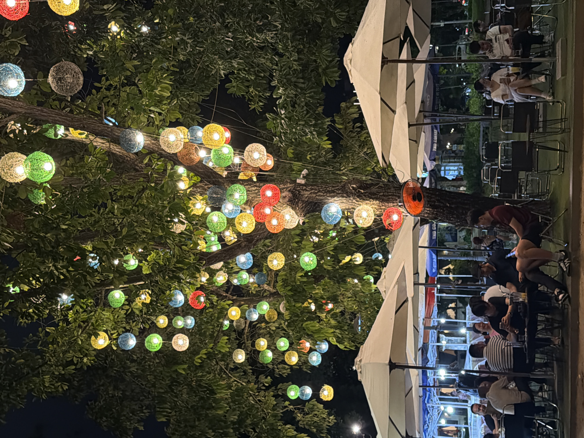 This is a nighttime, eye-level, full shot of an outdoor seating area under a large tree adorned with numerous colorful, spherical lanterns. The lanterns, in shades of green, yellow, blue, red, and white, are suspended at various heights from the tree's branches and emit a soft, warm glow.
In the foreground, several white umbrellas provide cover for tables and chairs. A group of people, appearing to be patrons of a cafe or restaurant, are seated at tables. Some are engaged in conversation, while others are looking at their phones or holding drinks. The individuals are dressed casually.
The environment suggests a relaxed, social setting, likely a cafe or outdoor dining establishment in a Vietnamese city. The presence of streetlights and other ambient lighting indicates an urban environment. The ground appears to be covered in artificial turf or a similar material. The overall atmosphere is inviting and festive due to the illuminated lanterns.
The location is identified as Thuận Hóa Ward, Vietnam.