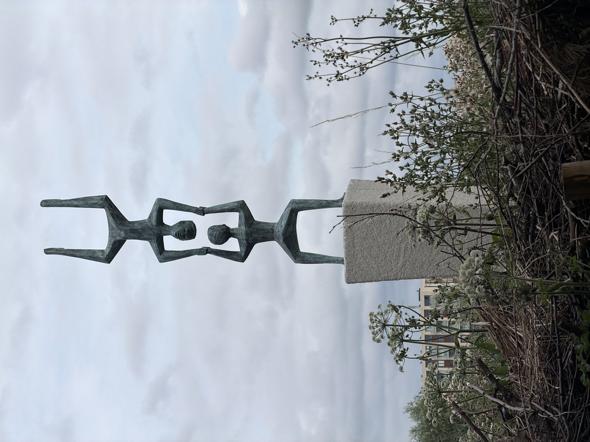 This is a full shot of a bronze sculpture of three stylized human figures stacked vertically, located outdoors in the city of Rijswijk, Netherlands. The sculpture is mounted on a rough-hewn stone pedestal. The bottom figure is standing, facing forward, with arms outstretched to the sides. The middle figure is standing on the shoulders of the bottom figure, facing forward, with arms also outstretched. The top figure is upside down, with its feet on the shoulders of the middle figure and its head pointing upwards. The figures are abstract and minimalist in design.
The background shows an overcast sky with scattered clouds. In the middle ground, there is a dense arrangement of dry, brown branches and twigs in the foreground, with green plants and white flowers growing around and in front of them. Behind the branches and plants, a light-colored building with multiple stories and visible windows is visible in the distance, partially obscured by the vegetation. The overall environment appears to be a somewhat unkempt, natural area within an urban setting.