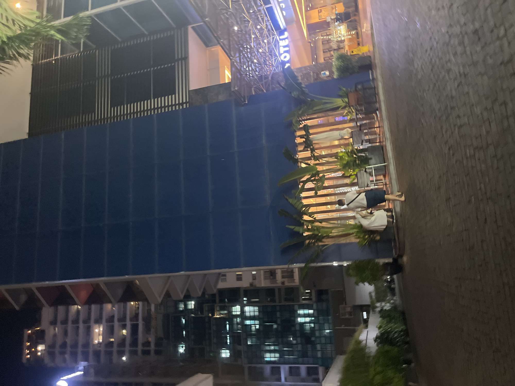 The image depicts a nighttime scene in Nha Trang, Vietnam, likely in a commercial or hotel district, given the presence of modern buildings. In the foreground, a brick-paved area serves as a pathway or entrance. Two individuals are visible: one person wearing a white dress and another in a white t-shirt and blue shorts, both walking away from the camera.
To the right, a large building with a dark blue facade dominates the midground, featuring a distinctive horizontal striped pattern of dark slats. Above this, a section of the building appears to be a glass facade with illuminated horizontal blinds. Further to the right, a canopy structure with intricate metal supports is illuminated from below, with the word "OTEL" visible in bright blue neon lettering, suggesting a hotel entrance or establishment.
In the background, a tall building with a glass facade is lit by numerous interior lights, revealing multiple floors and windows. Adjacent to this building, another taller structure with visible balconies and lit windows is present. Lush green foliage, including potted plants and trees, is strategically placed around the entrance area, adding a touch of nature to the urban environment. The overall atmosphere is illuminated by artificial lights, indicating the scene is taking place after sunset.