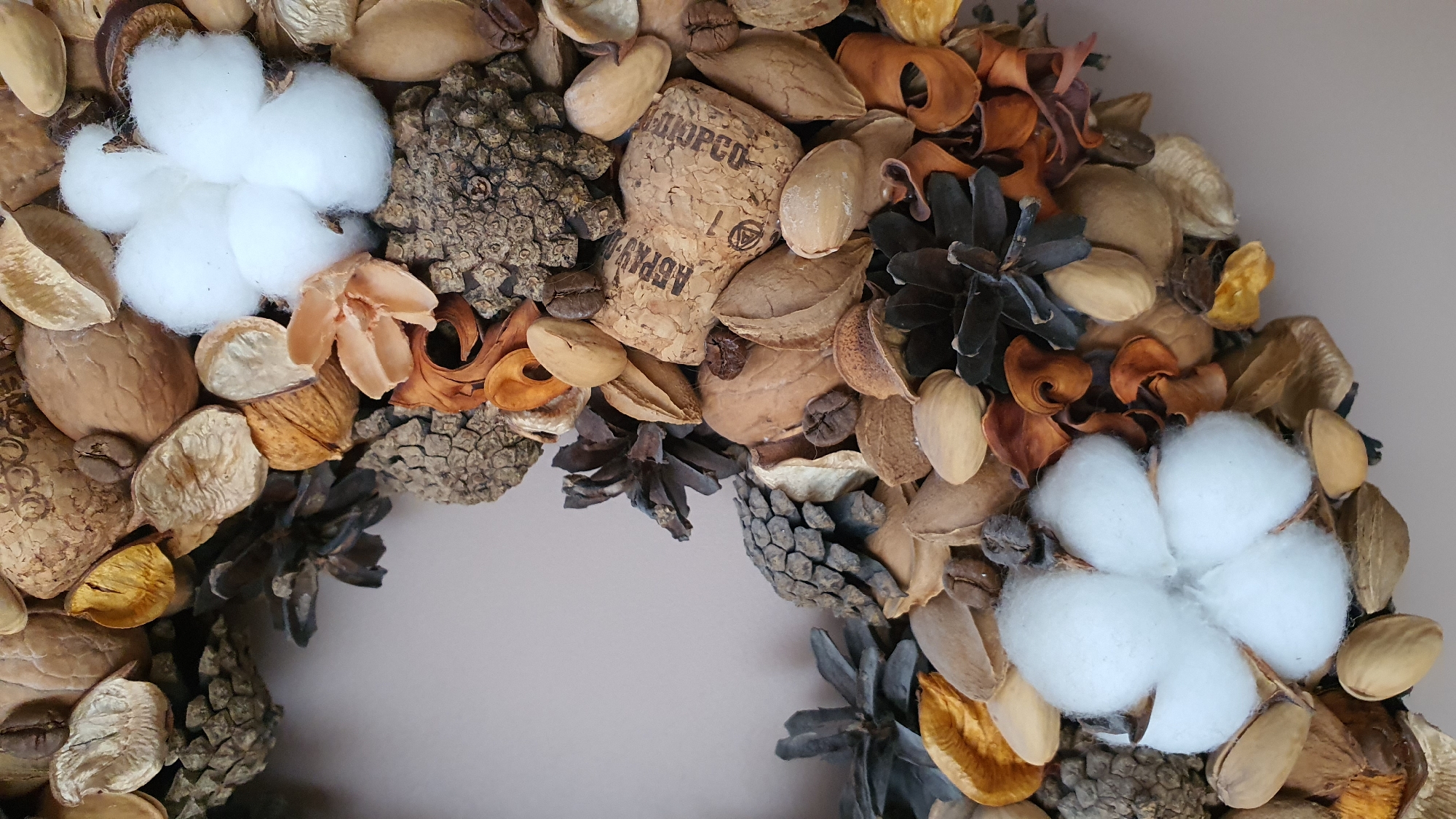 The image is a close-up, overhead view of a decorative wreath composed of natural materials such as nuts, pinecones, dried flowers, and cotton bolls. The wreath is circular and appears to be arranged on a flat, light-colored surface, possibly a table or wall. The lighting is soft and diffused, highlighting the textures and colors of the natural elements. There are no people or animals visible in the image. The background is a solid, neutral tone. This image does not provide any information about its location in the city of Krasnoyarsk, Russia, or any other geographical context.
