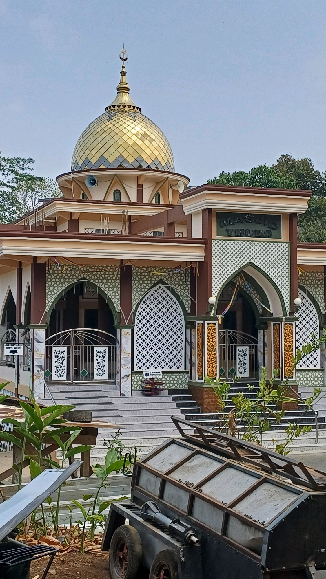 This is a full-shot, eye-level, outdoor photograph of a mosque in Pringsurat, Indonesia. The mosque features a large, golden dome with a crescent moon and star finial. The building is adorned with intricate geometric patterns in green and white. The entrance to the mosque is marked by an archway with the mosque's name, "MASJID THUIRIA," written above it in Arabic script. Greenery and trees are visible behind the mosque, and the sky is clear. In the foreground, there is a trailer with a metal structure on it, and some plants. The overall scene suggests a well-maintained place of worship.