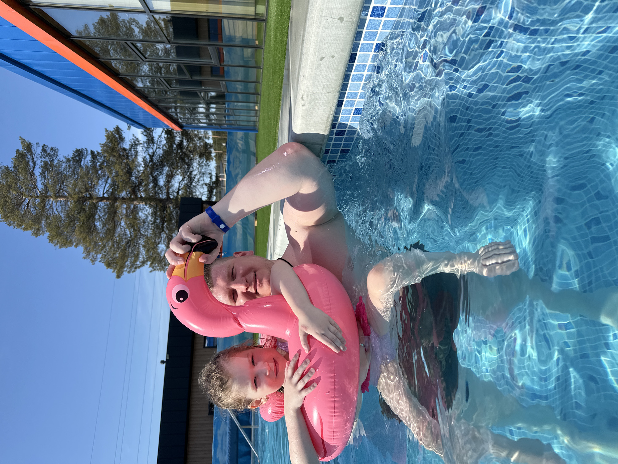 A family poses in a swimming pool, likely outdoors, located in Votkinsk, Russia. A fair-skinned adult male holds sunglasses while embracing a young girl with a pink inflatable flamingo. The adult and child are partially submerged in the blue-tiled pool. Reflected in the water are hints of other people swimming. The pool is edged with blue and white tiles and a concrete border. Beyond the pool's edge is a modern building with blue and orange siding and reflective windows, suggesting a hotel or recreation center. A grassy area is visible between the pool and the building. A clear blue sky is visible above.