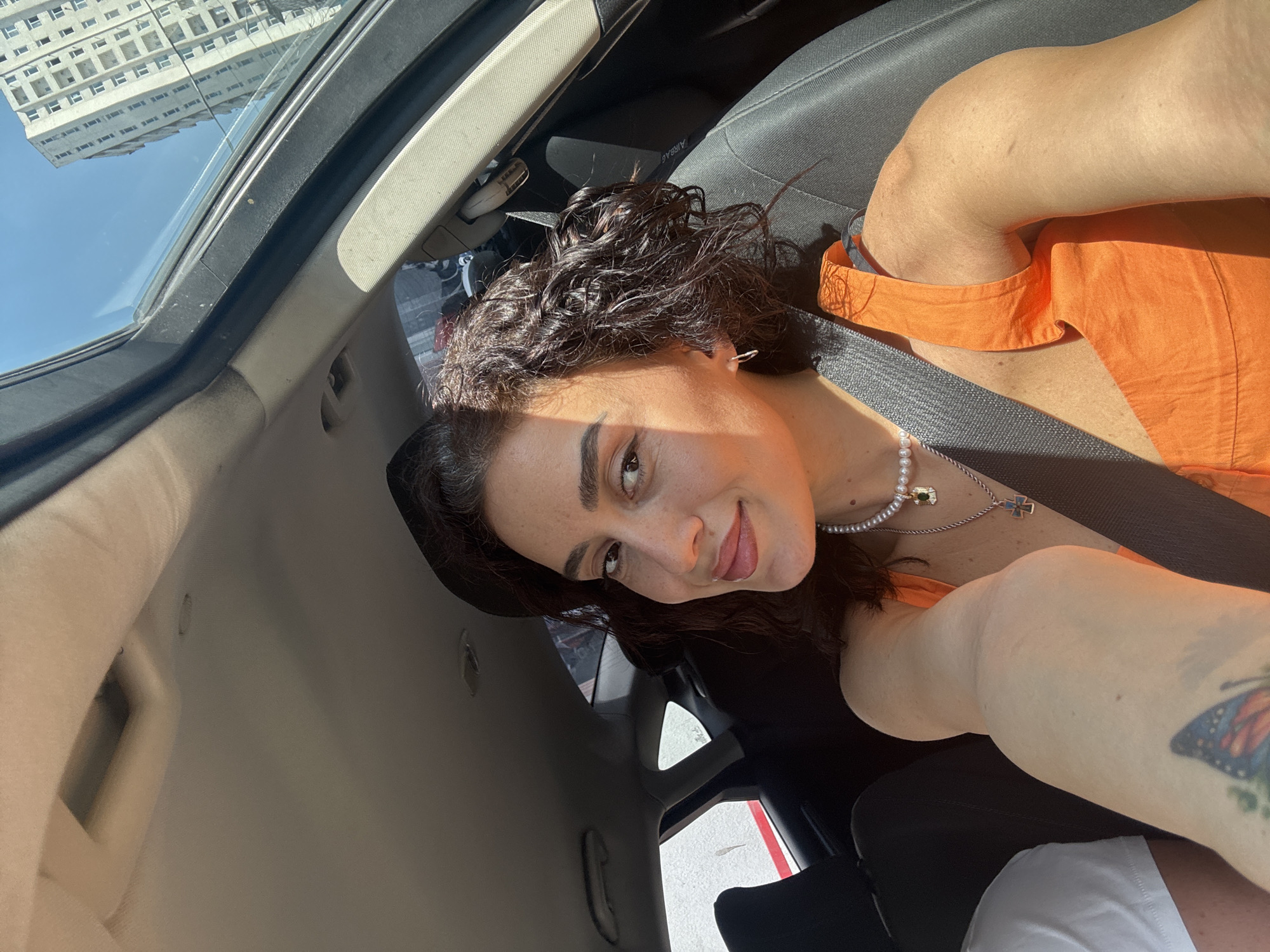 A young woman with curly dark hair is taking a selfie from the passenger seat of a car. She is wearing an orange sleeveless top and a pearl necklace with two pendants. A seatbelt is visible across her body. Sunlight illuminates her face and arm. Outside the car window, a tall, modern building and a clear blue sky are visible. The interior of the car, including the ceiling and seats, is also in view. The scene is consistent with being in the city of Tbilisi, Georgia, given the architecture of the building.