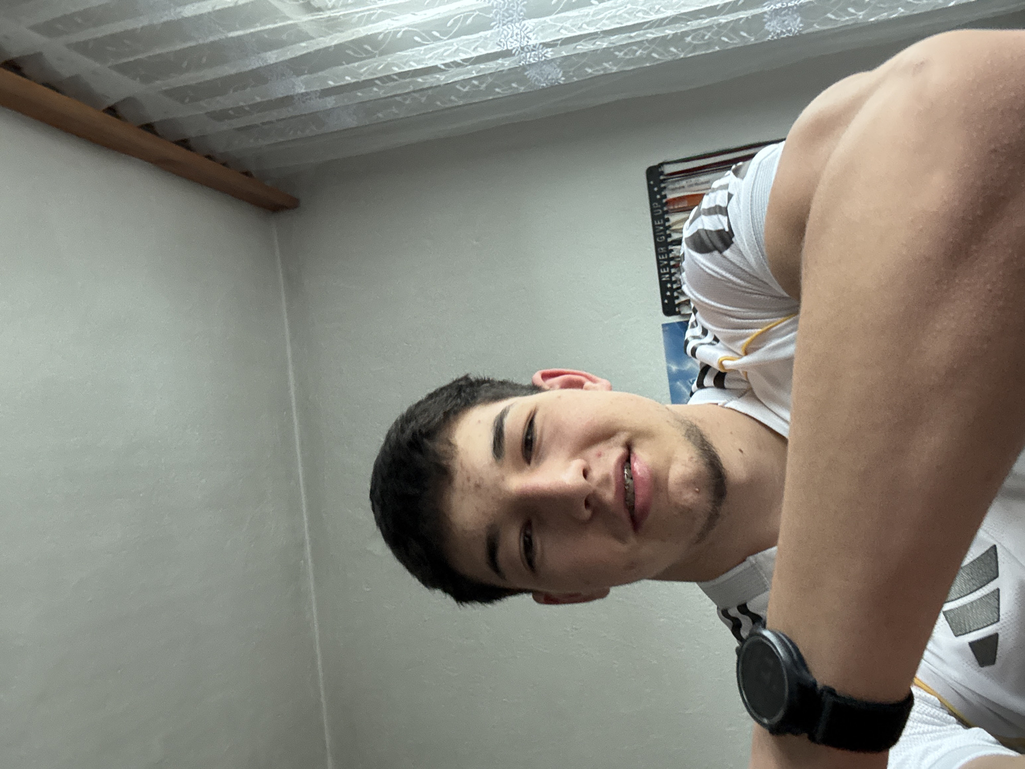 A young man is depicted in a close-up, upward-angled selfie, his head tilted to the left. He has dark hair, dark eyes, and a light complexion, displaying a slight smile that reveals braces on his upper teeth and light stubble on his chin. He is wearing a white t-shirt with black and grey striped patterns on the sleeves and shoulder, consistent with an athletic brand. A black smartwatch is prominent on his left wrist.
The setting is an indoor room characterized by light-colored walls. The ceiling, visible in the upper part of the frame, appears to be a suspended or paneled type with a subtle textured or patterned design. A wooden beam runs horizontally along the upper left corner of the room. To the right of the man's head, part of a dark notebook or binder is visible, leaning against a wall, with the text "NEVER GIVE UP" printed vertically on its spine. The lighting suggests an indoor environment, likely artificial. The overall expression of the man is casual and relaxed. While the image is contextualized to Zlatograd, Bulgaria, there are no specific visual elements within the photograph itself to identify this location.