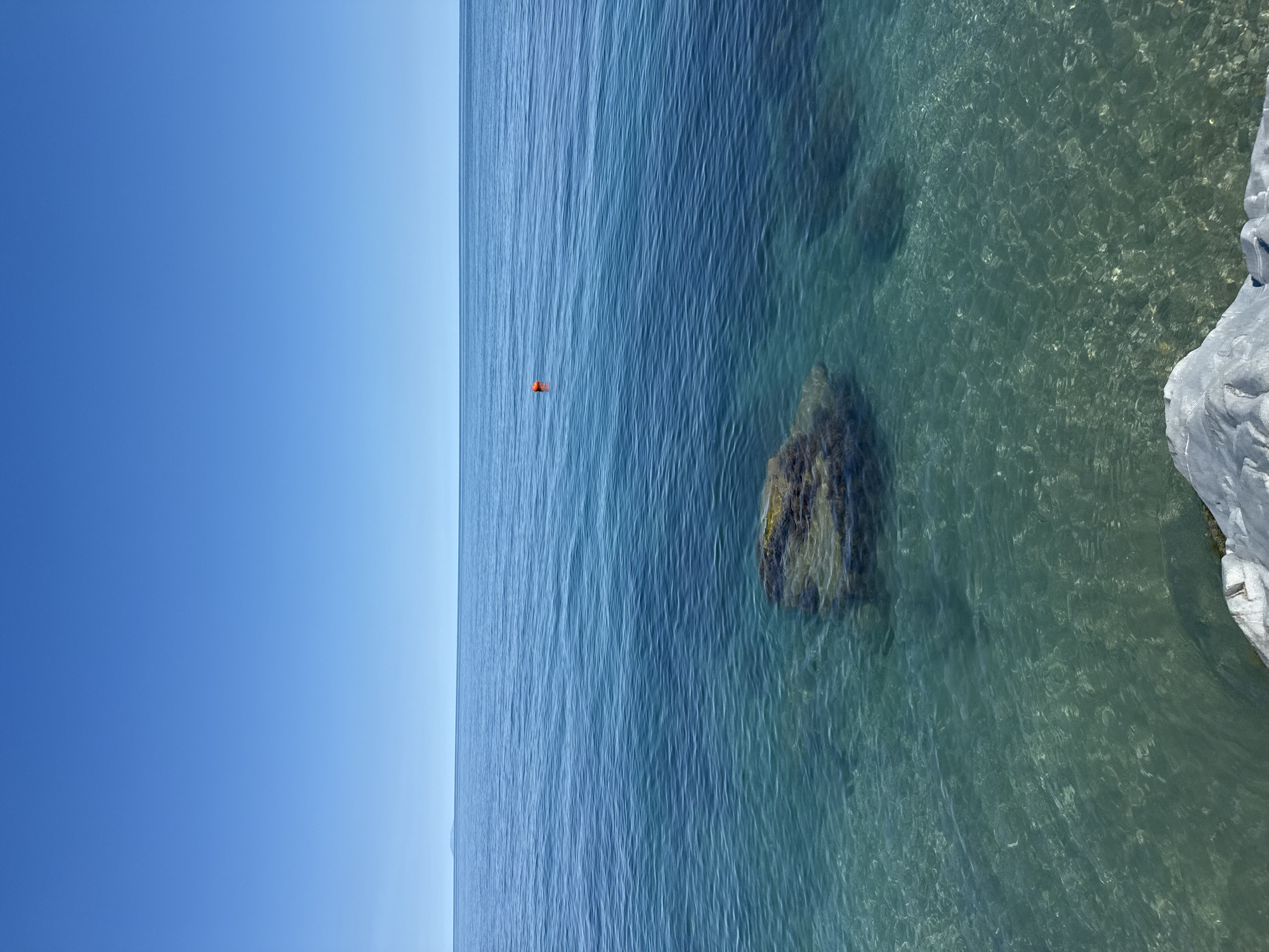 The image, when viewed in landscape orientation, captures a serene coastal scene dominated by a clear blue sky and calm waters. A sharp, level horizon line divides the expansive sky from the sea. The sky is a uniform, light azure, suggesting bright, sunny weather.
The ocean occupies the majority of the frame, displaying a gradient of blue hues. Further out, the water is a deep blue, transitioning to a clearer, turquoise nearer the shore. This transparency reveals dark, rocky formations beneath the surface. Gentle ripples create subtle patterns across the water's surface, indicating calm conditions.
In the middle distance, a small, bright orange object, possibly a buoy or a personal floatation device, floats distinctly on the water. To the far right, a segment of a light-colored, textured rock formation, likely part of a cliff or shoreline, frames the scene. No people or text are visible in the image. The overall impression is one of peaceful natural beauty.