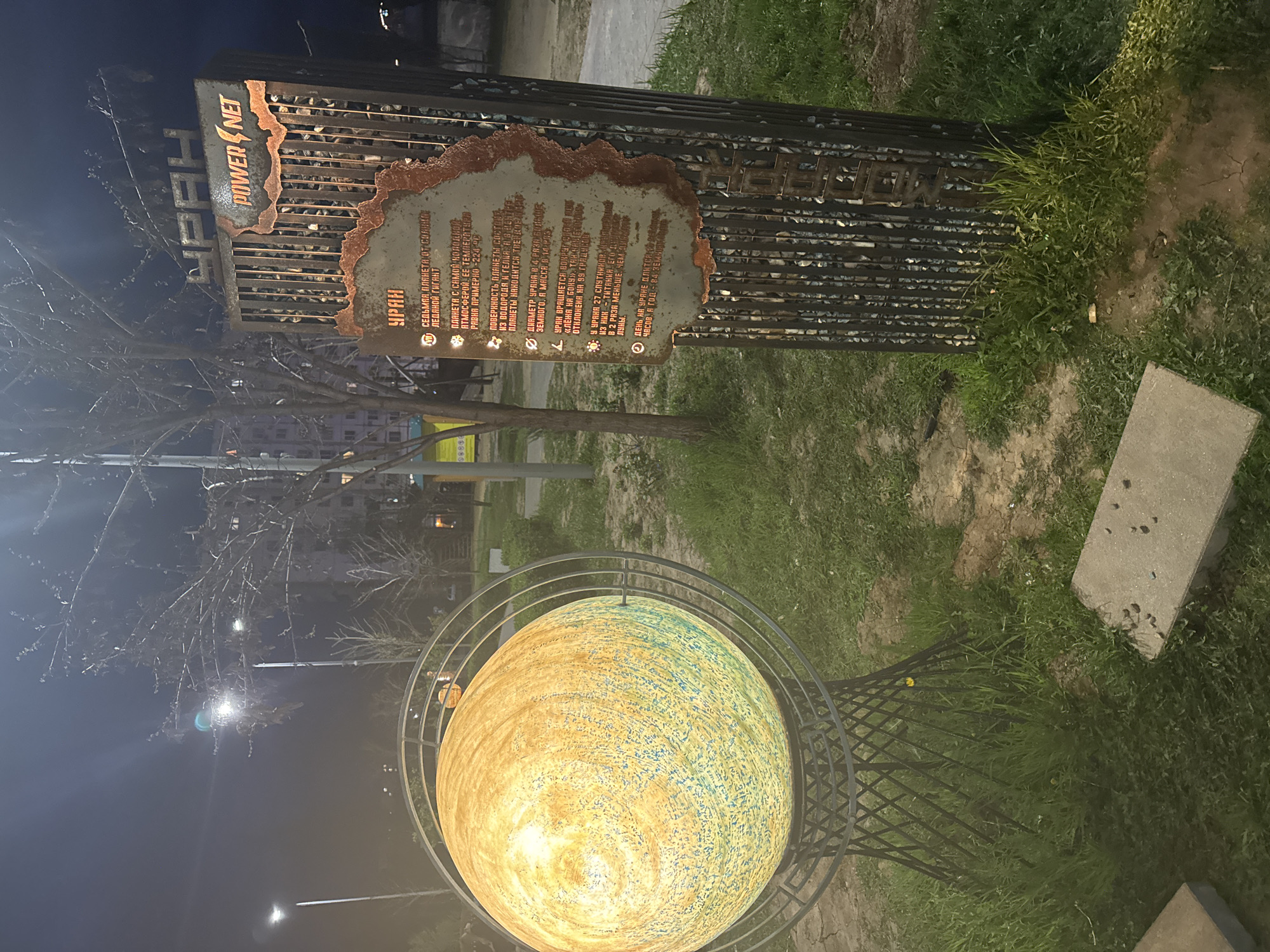 The image shows a nighttime shot of an outdoor display, likely in Volzhsky, Russia, as indicated by the Cyrillic text on the sign. A stylized metallic structure reads "POWER NET" above an information plaque about the planet Uranus. Below this is a spherical representation of Uranus surrounded by a circular metal frame. The sphere is painted with shades of yellow and teal to resemble the planet. Grass and a paving stone are visible around the base. Bare tree branches are in the background, and streetlights illuminate the scene.