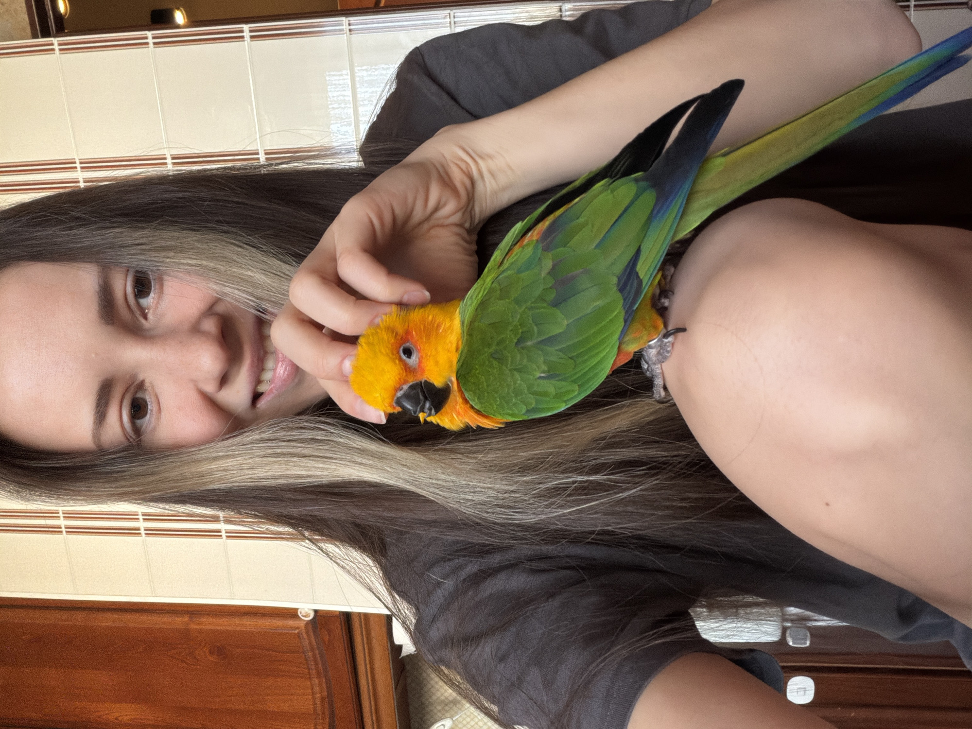 A woman is holding a colorful bird, likely a sun conure, against her arm. She is smiling at the camera. The bird is perched on her arm, and she is gently touching its head with her hand. The background includes a tiled wall and a wooden cabinet. There is no evidence to suggest that the image was taken in Khimki, Russia.