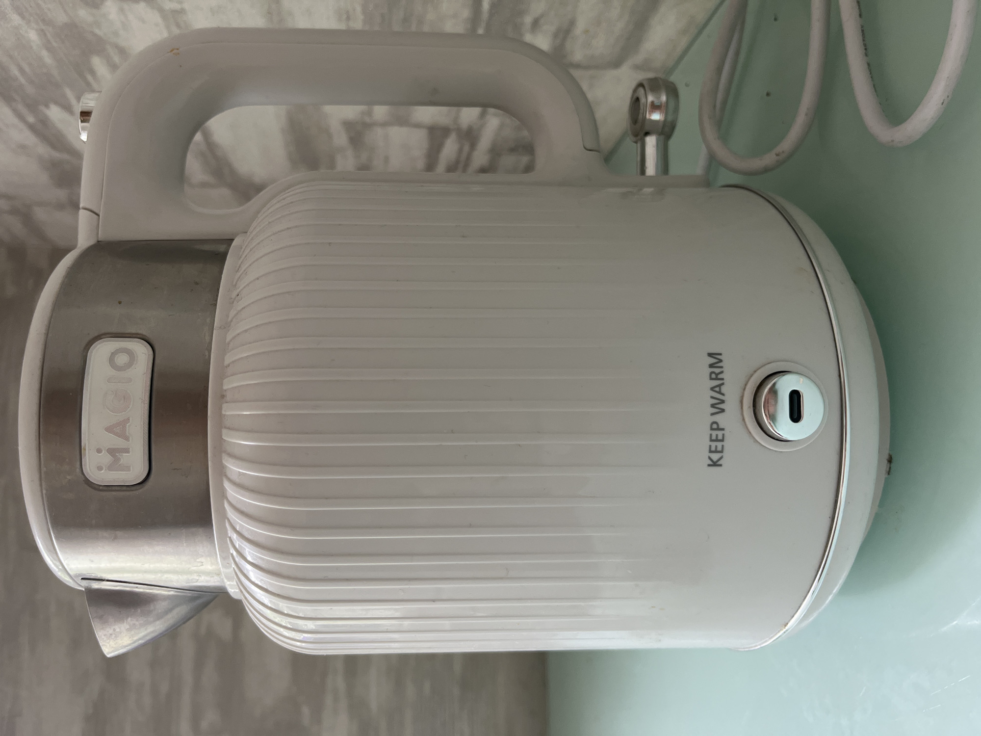 A white electric kettle with vertical ridges sits on a light green surface. The kettle has a silver lid with a logo that reads "MAGIO". A white handle extends from the right side of the kettle, and a silver spout is visible on the left. Below the spout, the text "KEEP WARM" is printed. A silver power button with a USB-C port is located at the base of the kettle. A white power cord is coiled next to the kettle. The background consists of a grey, textured wall. The scene is indoors. The location is not specified in the image or the provided crops.