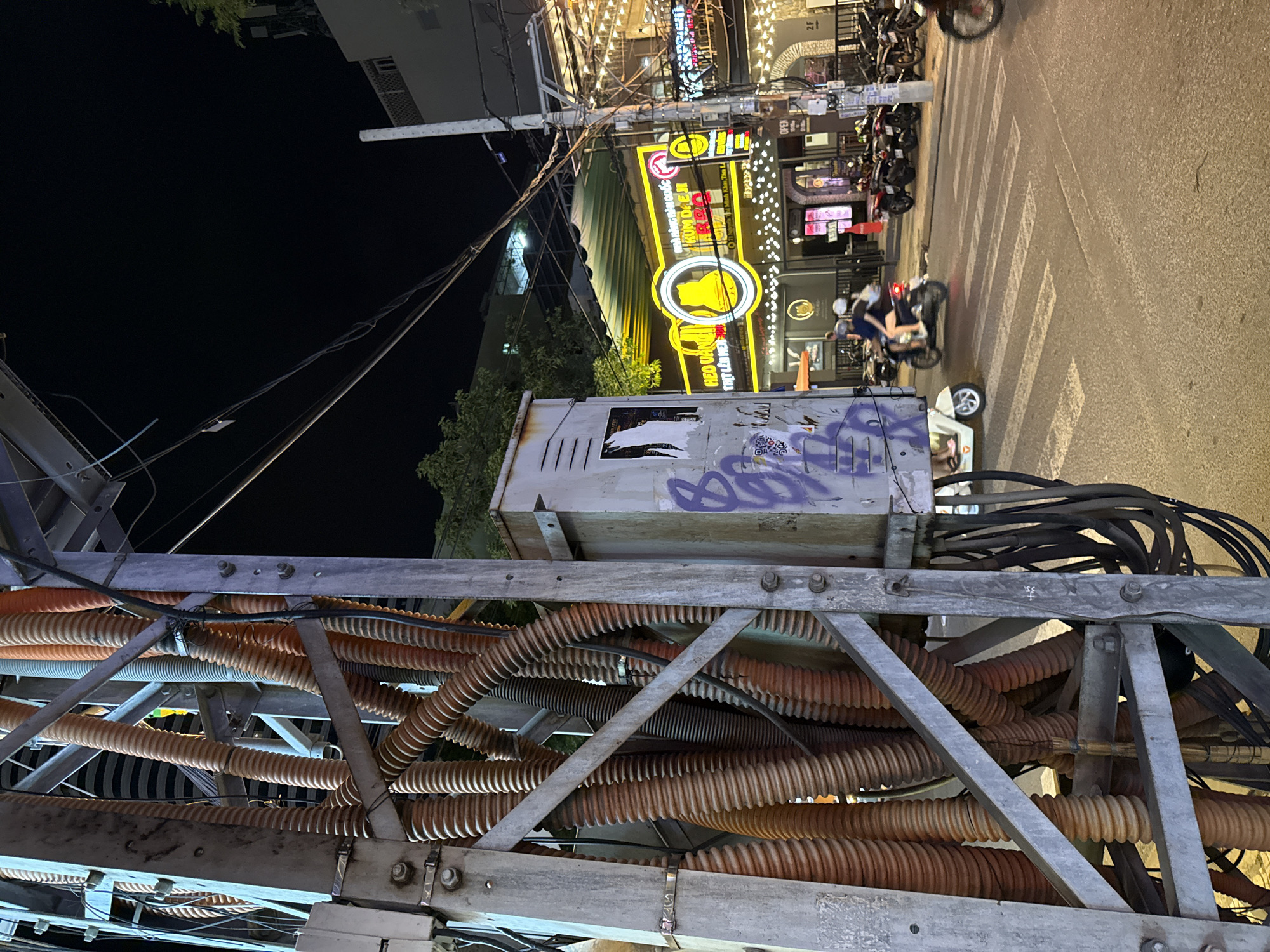 The image depicts a street scene at night in Nha Trang, Vietnam. In the foreground, a metal pole supporting a bundle of orange corrugated tubes and electrical wires dominates the left side of the frame. A gray electrical box with graffiti on it is attached to the pole. The street is paved, with a crosswalk marked by white stripes. Several motorcycles are visible on the street.
In the background, a building with neon signage is illuminated. The sign, in yellow and red neon, features a cat logo and Vietnamese text. The building appears to be a restaurant or establishment, with strings of small lights decorating its facade. Other buildings and trees are visible in the distance. The sky is dark, indicating nighttime. Two people are riding on a scooter, and another person is on a white scooter. A person on a motorcycle is blurred in the far right of the frame, suggesting movement. The overall scene suggests a busy urban environment at night.