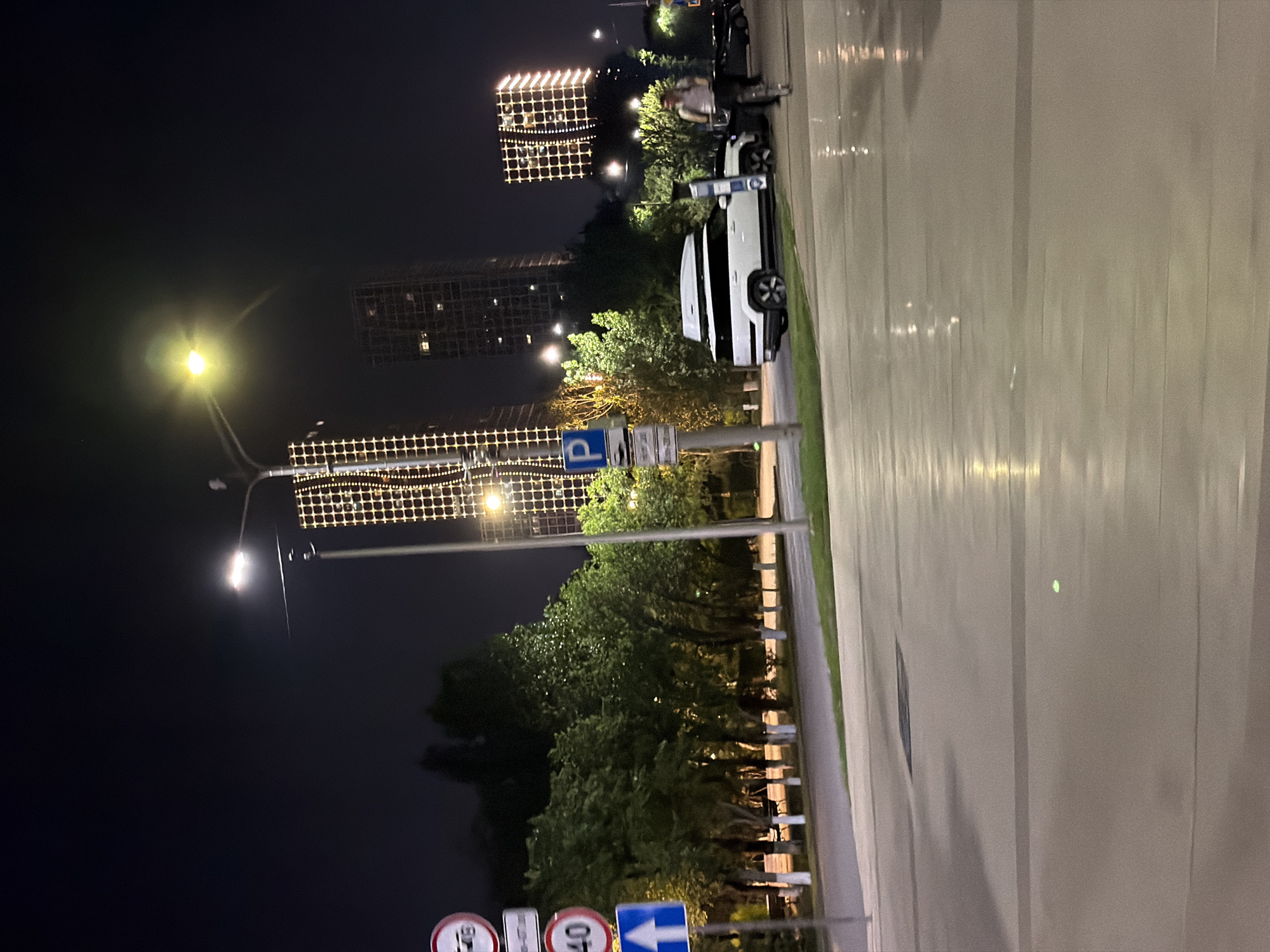The image depicts a nighttime scene on a street in Astana, Kazakhstan. In the foreground, a wet asphalt road reflects the lights from the surroundings. A white SUV with a roof box is parked on the right side of the street. Further down the street, a person on a scooter is visible. Behind the parked vehicle and scooter, trees line the street, and tall buildings with illuminated grid-like patterns of lights are prominent in the background. Several traffic signs are also visible on the left side of the image, including a speed limit sign indicating 40 and other regulatory signs. Streetlights illuminate the area, casting a glow on the road and surrounding elements. The overall atmosphere is urban and nocturnal.