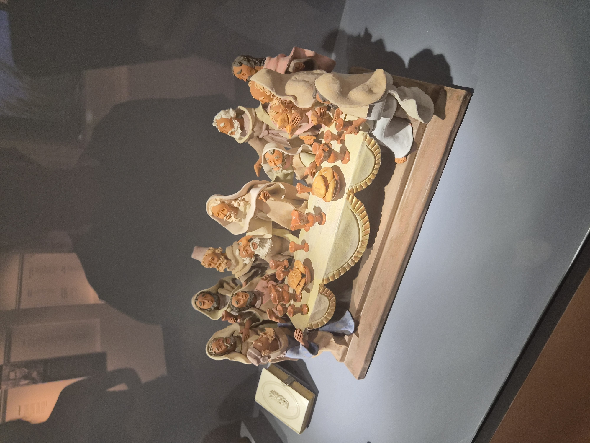 The image shows a religious ceramic sculpture depicting the Last Supper. The sculpture is set on a wooden base, which sits on a grey surface. Thirteen figures, dressed in robes, are gathered around a long table laden with bread and goblets. The figures appear to be engaged in conversation or communion. A book or tablet with intricate carvings is placed to the left of the table. The background is dimly lit with shadows suggesting the presence of people or other exhibits. This sculpture is located at the Museu de Artes Decorativas in Seia, Portugal.
