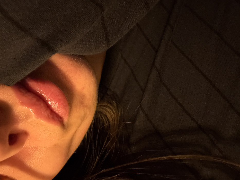 A close-up, slightly tilted view of a person's face partially obscured by dark fabric. The nose and glossy lips are visible, bathed in warm light. Strands of hair peek out from beneath the fabric, which features subtle vertical lines. The image creates a sense of intimacy, with the focus primarily on the soft texture and light reflecting on the face.