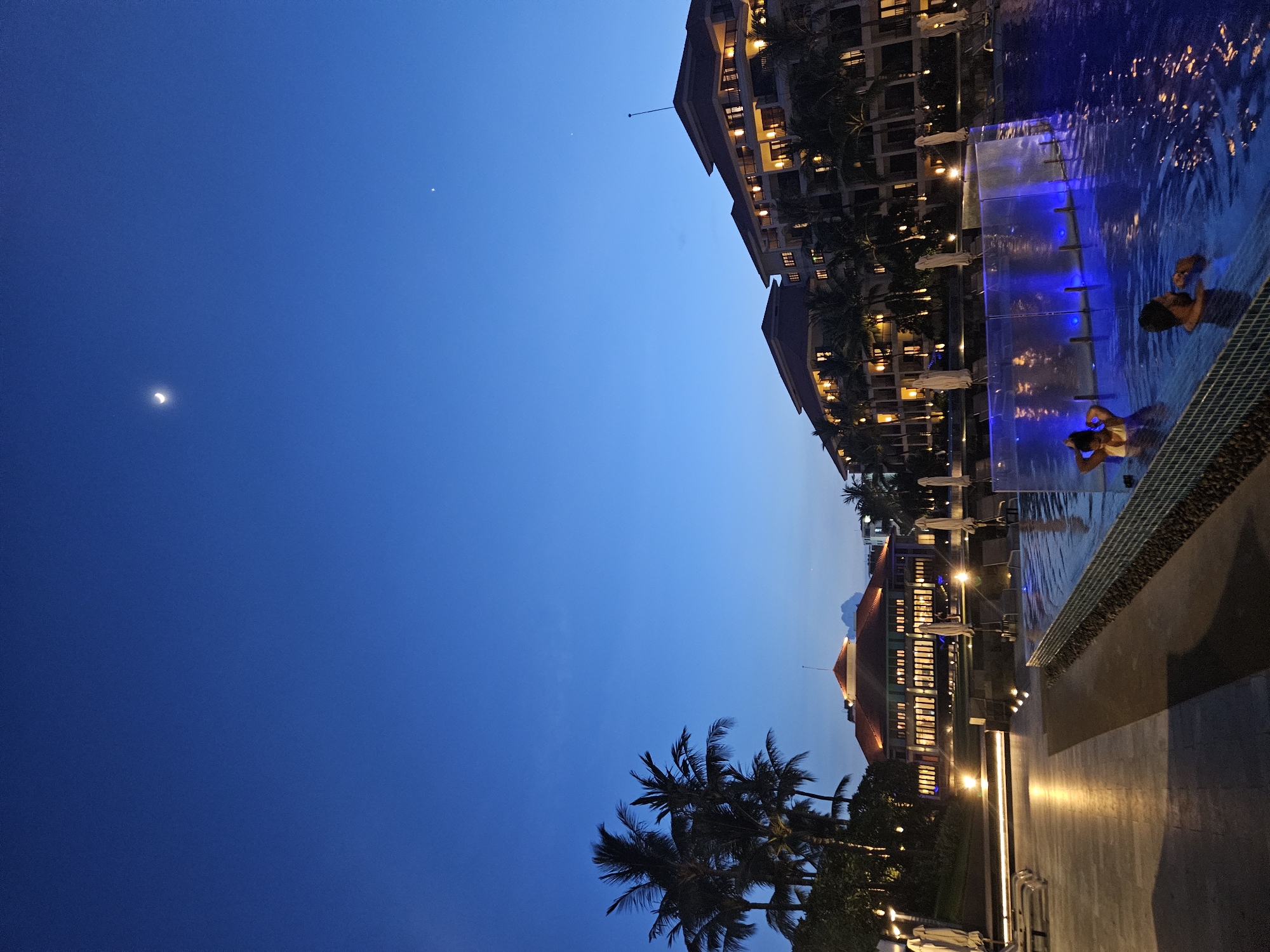 The image depicts a twilight scene at a resort in Da Nang, Vietnam. The sky is a deep blue, transitioning from the day's light. A crescent moon and a distant star are visible in the upper left quadrant.
The foreground features a swimming pool with illuminated blue lights, creating a vibrant glow in the water. Two individuals are partially submerged in the pool, appearing relaxed as they float or sit in the water. One person's head and shoulders are visible, looking towards the camera, while another's hands are resting on their head.
To the left of the pool, a large resort building is visible. It is multi-storied with numerous windows, many of which are illuminated by warm yellow light, suggesting occupancy and activity within. Palm trees are scattered around the building and the pool area, adding to the tropical ambiance. The ground around the pool is paved with dark tiles, and there are lounge chairs and umbrellas set up near the pool's edge.
The time of day is evening, indicated by the twilight sky and the artificial lighting of the resort and pool. The weather appears clear, with no clouds visible. The overall mood of the image is tranquil and inviting, capturing the relaxing atmosphere of a resort evening. No text is visible in the image.