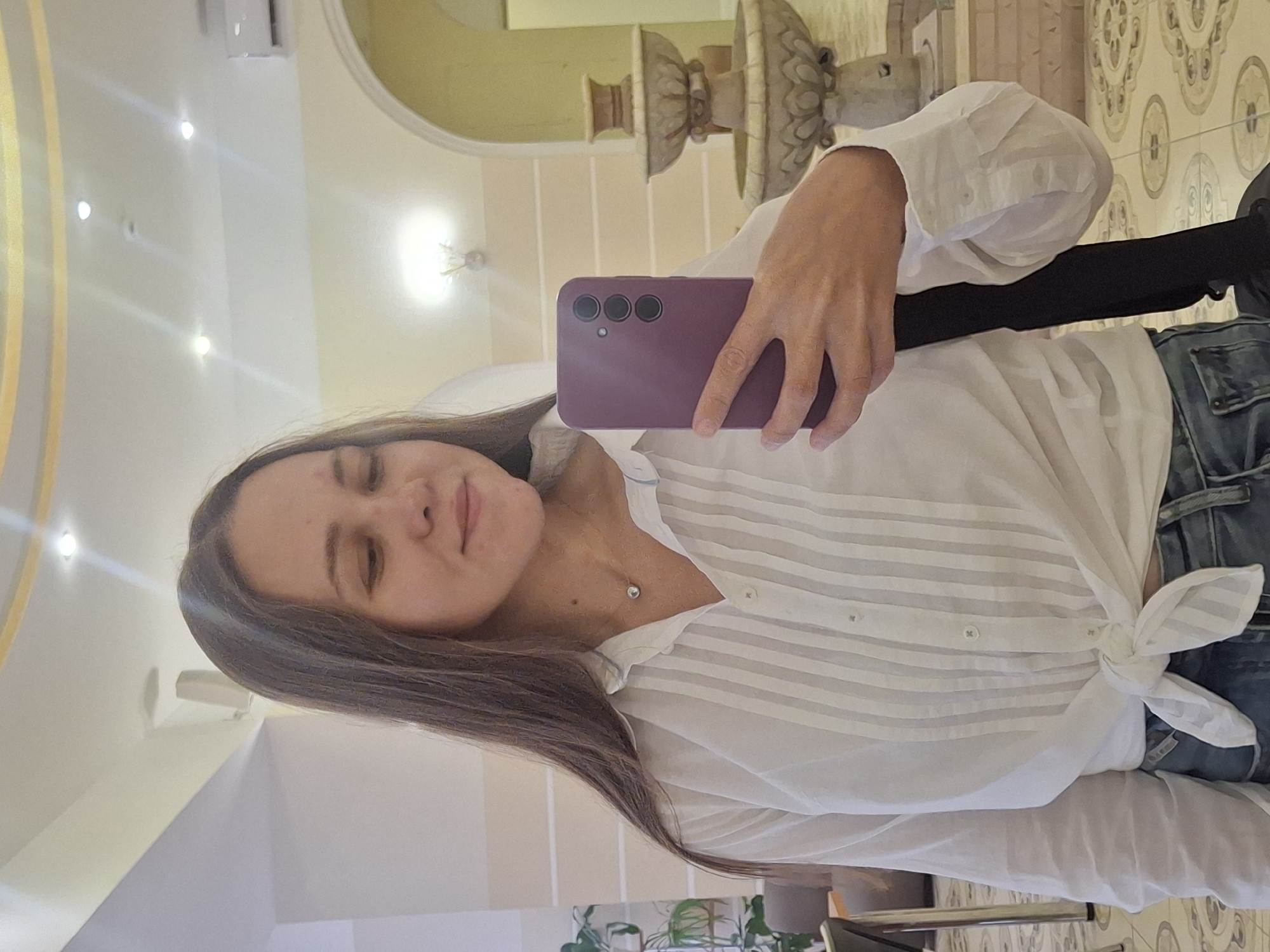 A young woman with long brown hair takes a selfie in a hotel lobby. She is wearing a white button-down shirt tied at the waist and blue jeans. Her phone, with a purple case, is visible in her right hand. In the background, a tiered fountain and a wall with arched doorways suggest an interior setting. The floor has a patterned tile design, and the ceiling features recessed lighting. The image does not provide any specific visual cues to pinpoint its location within the city of Chaikovsky, Russia.