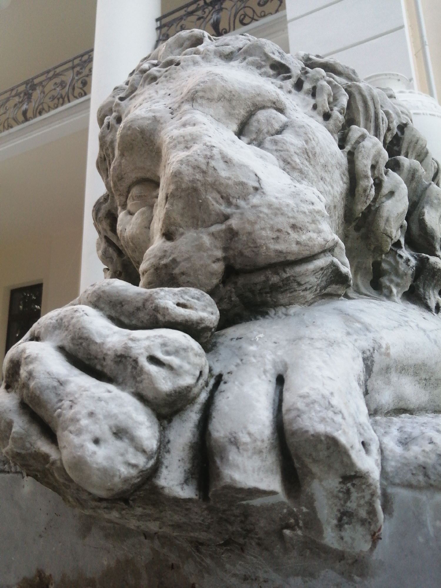 The image displays a close-up, low-angle shot of a weathered stone sculpture of a lion's head. The sculpture is intricately detailed, showing rough textures and some erosion, particularly on the mane and facial features. The lion appears to be resting its paws or head on a surface. In the background, out of focus, is a building with architectural elements, including a balcony with a decorative railing, suggesting an urban or historical setting. The lighting is diffused, creating soft shadows.
This particular sculpture is part of the lion statues located in the central park of Slonim, Belarus, a significant landmark within the city.
