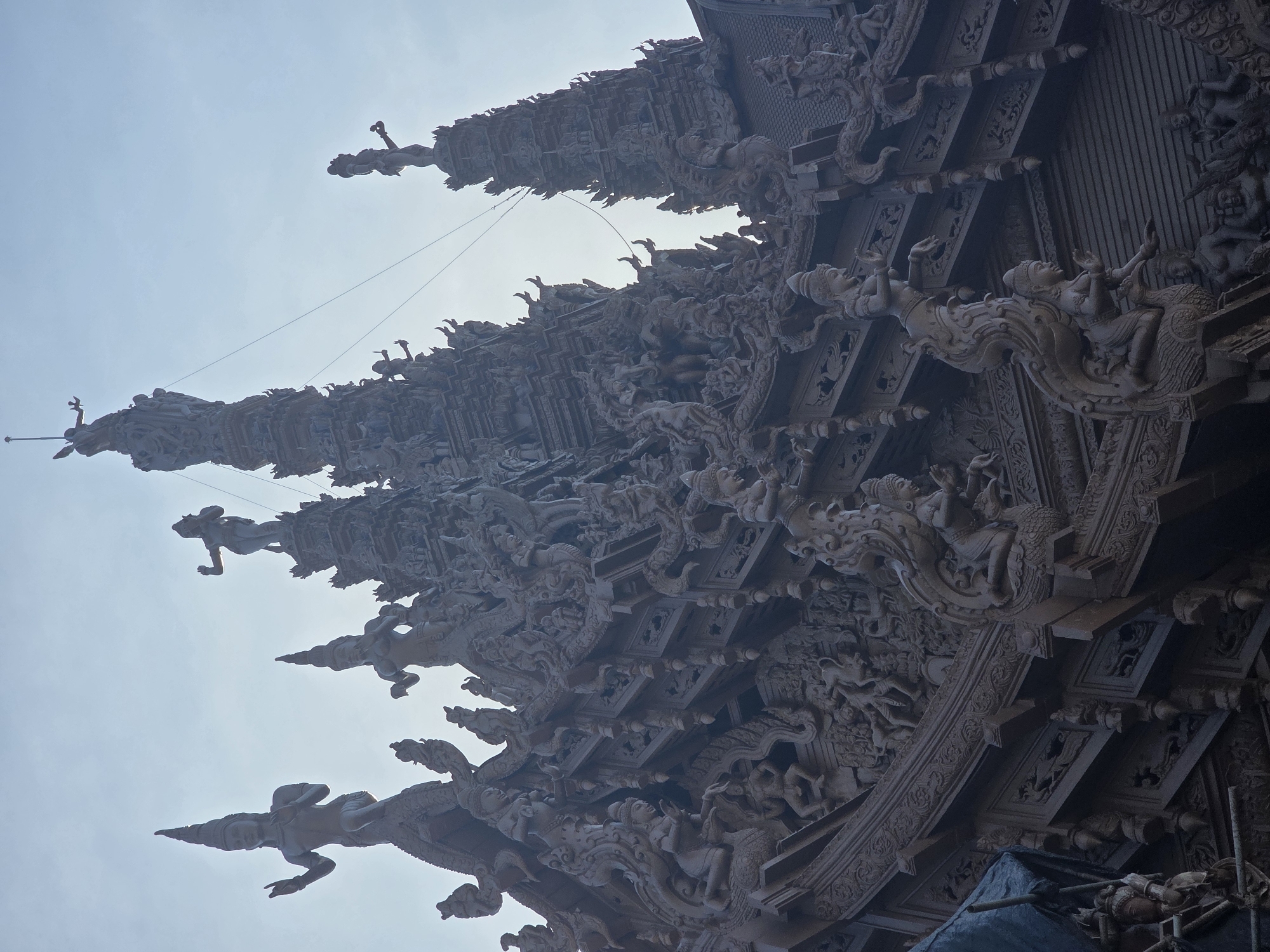 The image displays an intricate, ornate wooden structure, likely a temple or pavilion, set against a bright, hazy sky that suggests a daytime setting, possibly early morning or late afternoon due to the diffused light. The structure is adorned with numerous carved figures, predominantly human-like in form, engaged in various poses and scenes. These carvings are densely packed, covering the surfaces of the building with detailed reliefs and sculptures.
The style of the carvings appears to be traditional Southeast Asian, possibly Thai, given the location context of Pattaya City, Thailand. The figures exhibit flowing drapery, elaborate headdresses, and dynamic postures, some appearing to be in mid-leap or in reverent poses. Many of these figures are situated along the edges and eaves of the structure, resembling decorative elements or possibly depicting mythological scenes or deities.
The overall impression is one of awe-inspiring craftsmanship and spiritual devotion. The scale of the carvings suggests a significant monument. The hazy sky and the muted tones of the wood, combined with the dense, detailed carvings, create a somewhat ethereal atmosphere. There are no visible signs of people actively engaged in construction or maintenance, apart from some scaffolding and tarpaulin visible at the very bottom right of the frame, suggesting ongoing work or preservation efforts. No visible text is present in the image. The image is oriented vertically, with the structure appearing to extend upwards towards the sky.