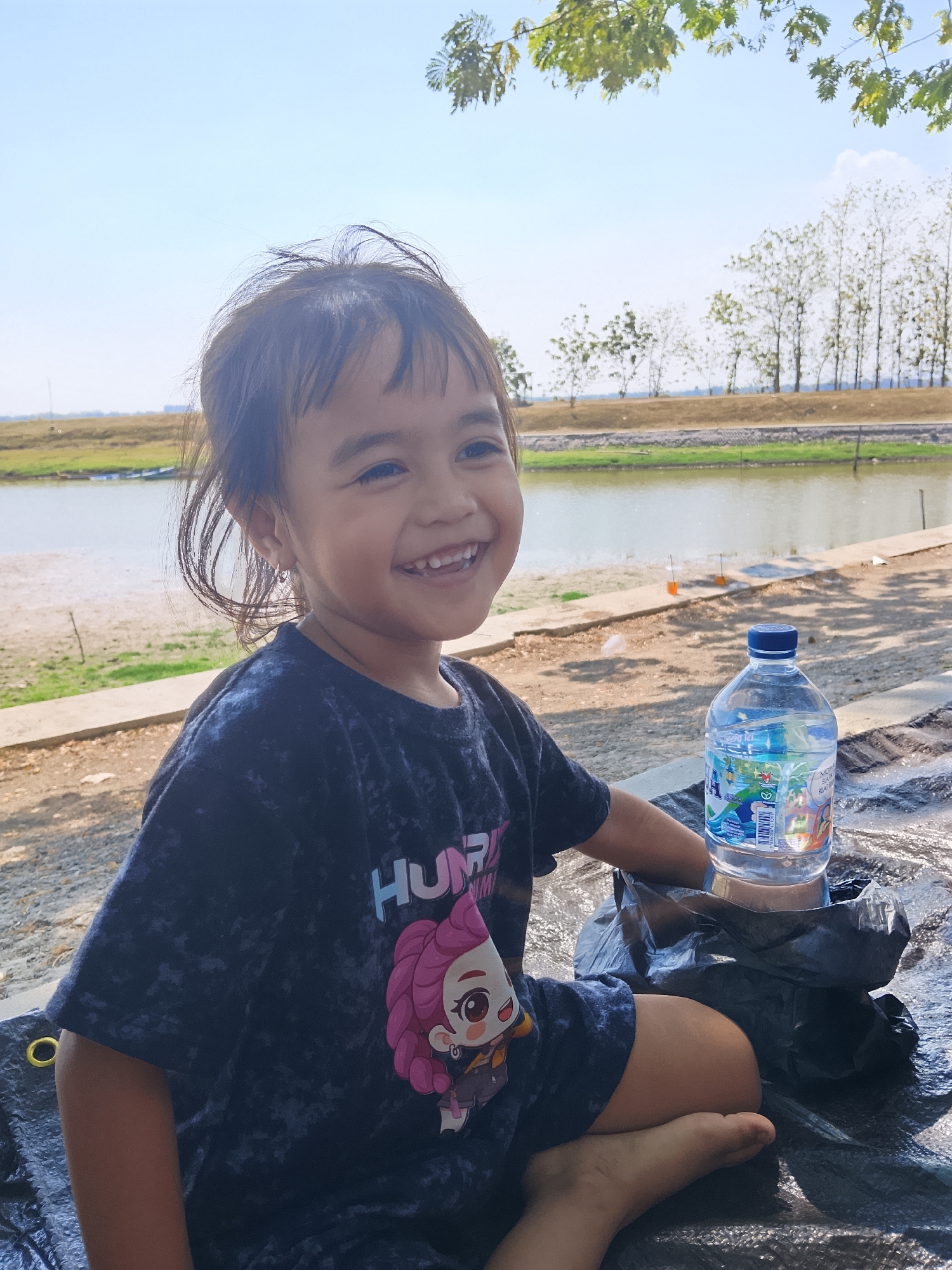 A young girl with dark hair and a black t-shirt with a cartoon character on it is sitting outdoors by a body of water. She is smiling and looking to her right. In front of her is a plastic bottle of water and a black bag. The background shows a tree-lined shore and a blue sky. The scene appears to be in the city of Ngadrin, Indonesia.