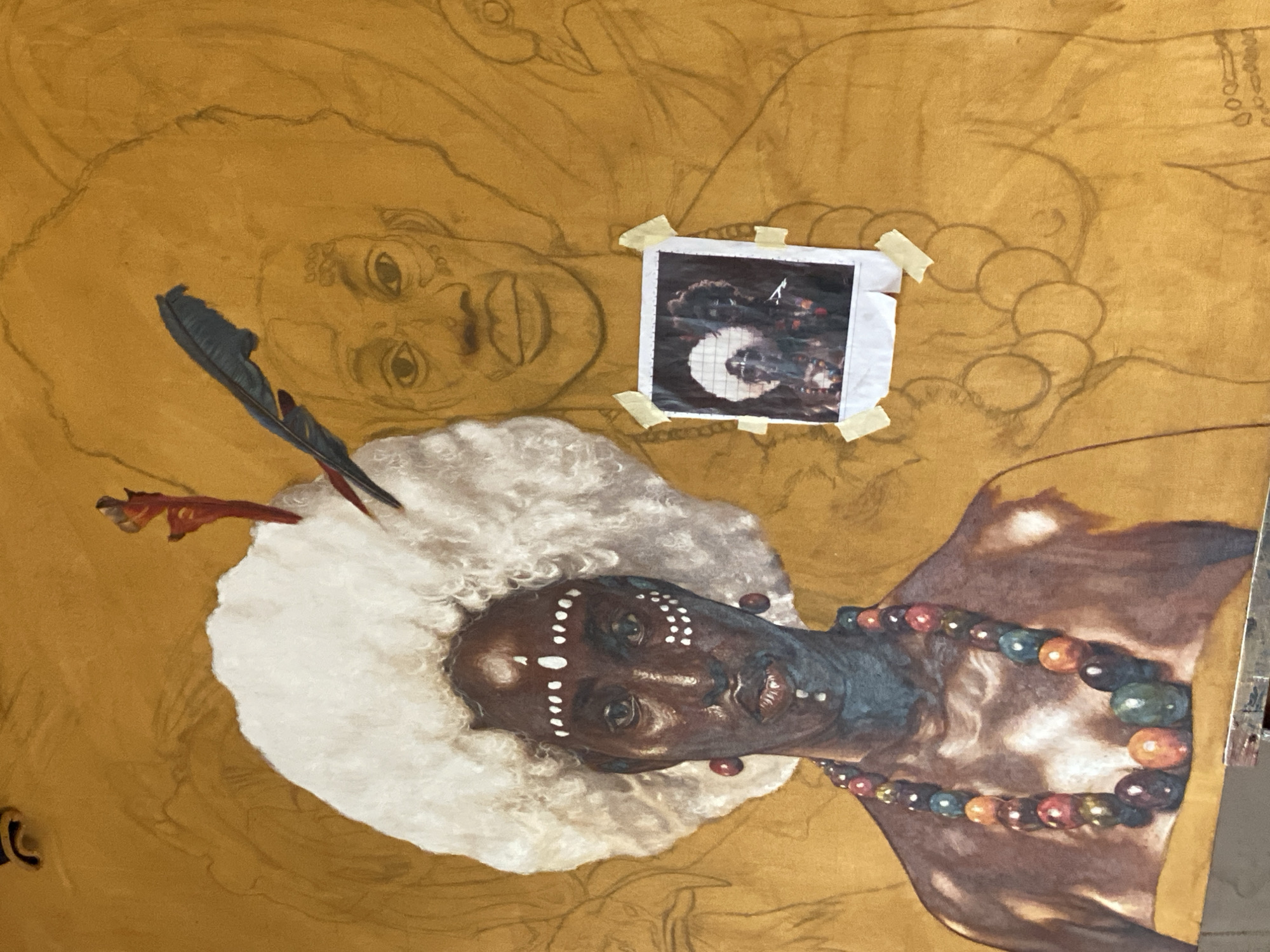 The image shows an unfinished painting on a gold-toned canvas. The central figure is a person with dark skin, adorned with white dots on their face and a large white headpiece with a dark feather. They wear a colorful beaded necklace. Above the figure is a pencil sketch of another person. A small, square photograph divided by a grid is taped to the canvas, near the sketched figure. The painting may be taking place in Ado, Nigeria. The subjects include people, the painting on canvas, and the photograph. Actions include the artist painting the figure. Interactions include the artist using the photograph as a reference. The environment appears to be an art studio or workshop.