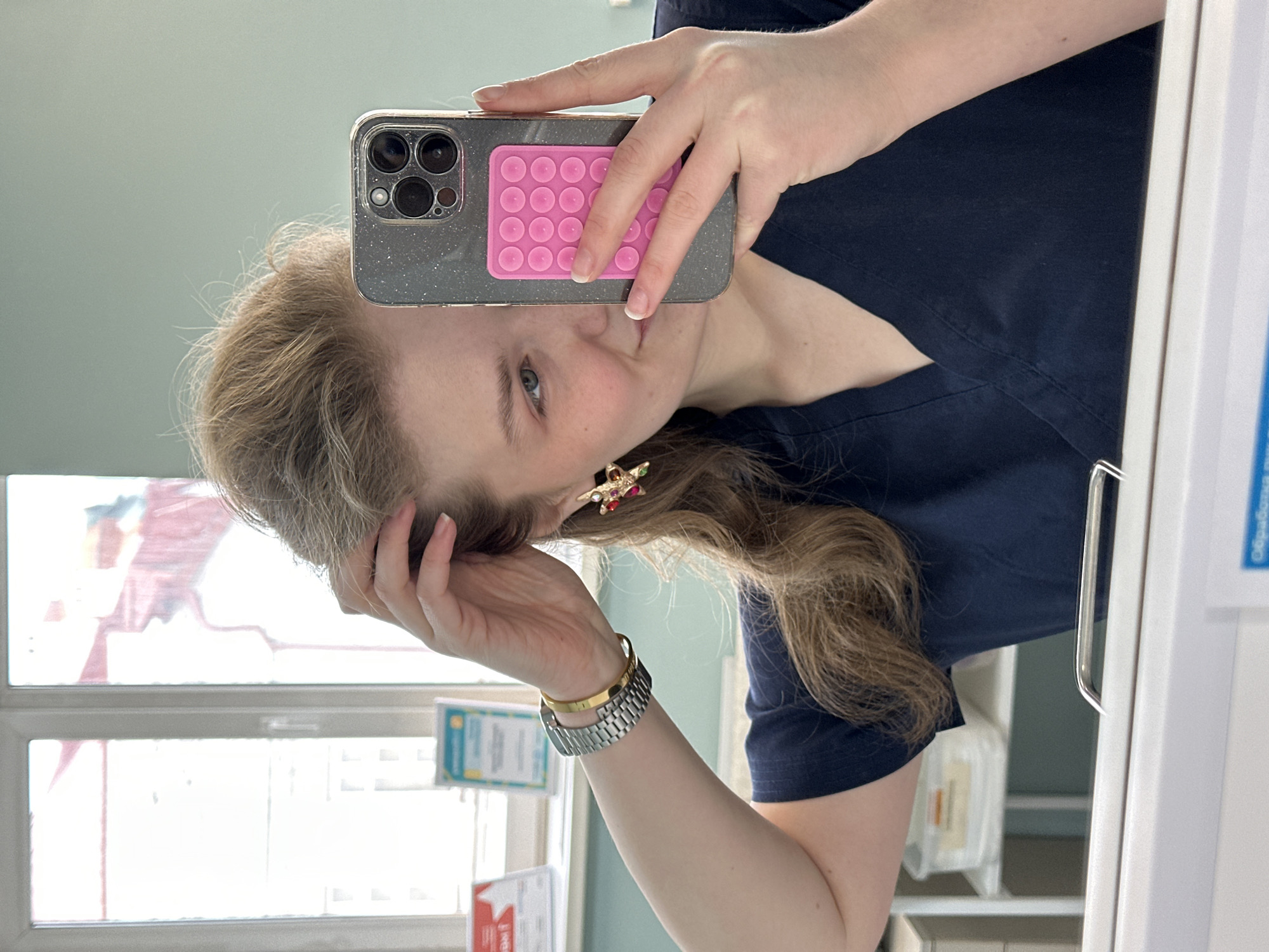 The image shows a woman taking a selfie in what appears to be an indoor environment. She is holding a smartphone with a pink pop-it attached to the back. The woman has fair skin, blue eyes, and light brown hair. She is wearing a dark blue V-neck top. On her wrist, she has a silver watch and a gold bracelet. She is touching her hair with her other hand.
Behind her, there is a window with a white frame and multiple panes. Various documents are posted near the window. To her right, there's a white cabinet or refrigerator with a handle. Based on the reflection of a red flag with white text in the window, and a sign near the window, it is likely that this photograph was taken in Kazan, Russia. The precise location is not clear from the image but appears to be some kind of office or medical setting.