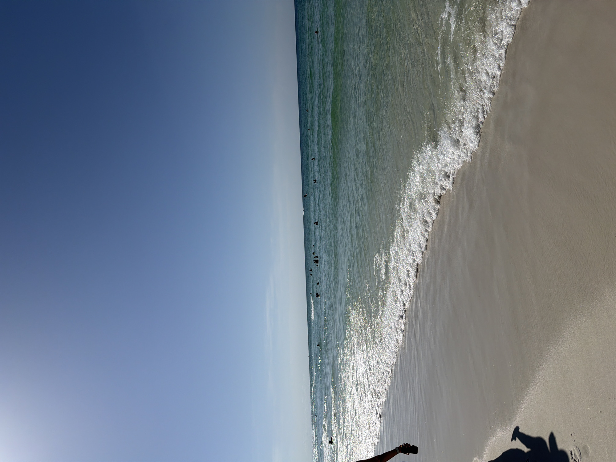 A vertical shot of a beach in Destin, Florida, shows a bright, clear blue sky dominating the upper half of the frame. The ocean, a vibrant turquoise, occupies the middle section, with gentle waves lapping at the shore. Several people are visible in the water, swimming and enjoying the sea. A white, foamy wave line extends across the bottom of the ocean portion, meeting the light tan sand of the beach. On the left side of the image, a person's shadow is cast on the sand, suggesting the sun is behind them. The overall scene conveys a tranquil beach day.
