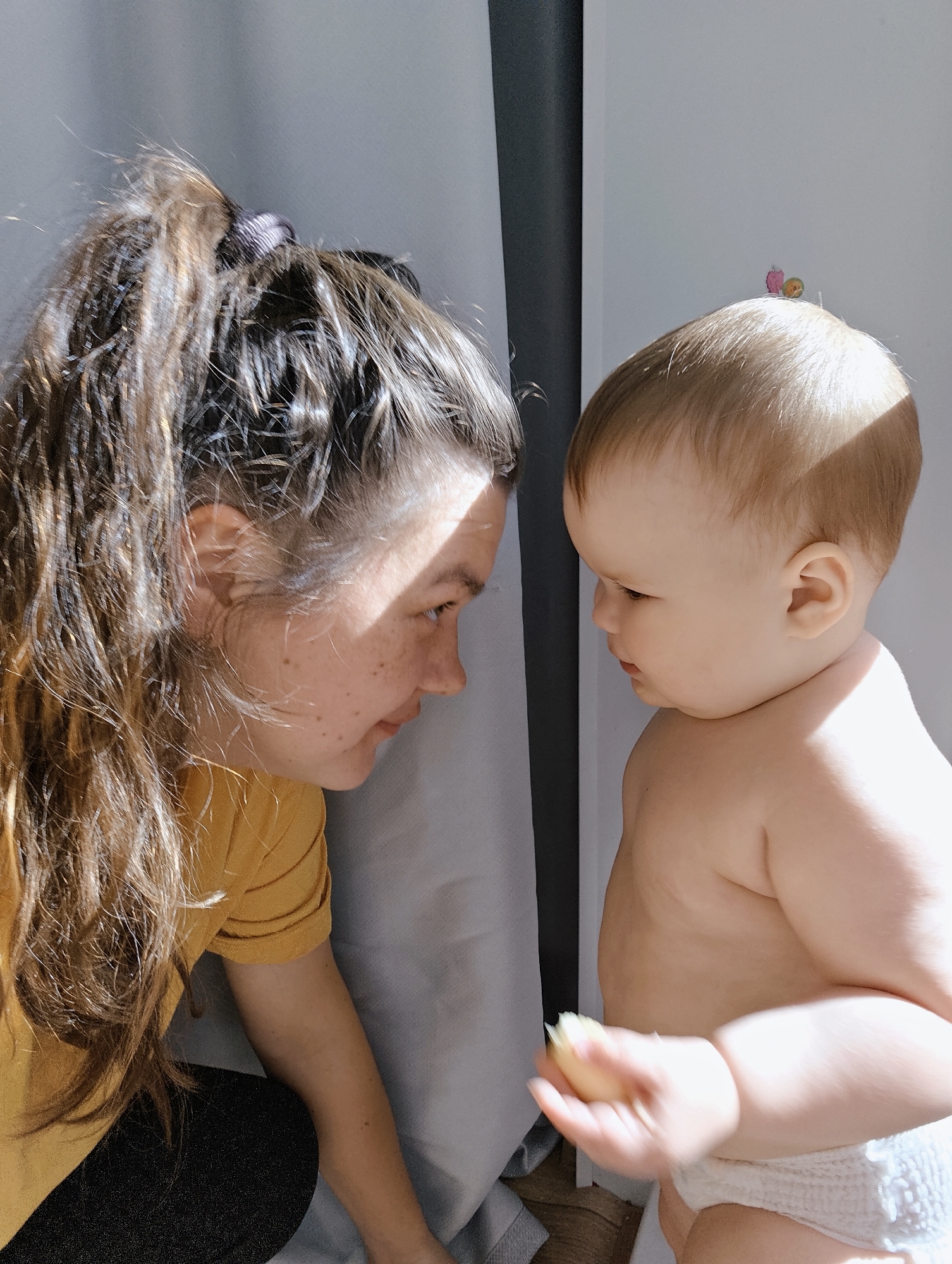A woman and a baby are facing each other indoors. The woman has long, brown hair in a high ponytail, wearing a yellow shirt and dark pants. Her face is close to the baby's, and she appears to be interacting with the baby. The baby is fair-skinned, wearing a diaper, and holding a piece of food in their hand. They are standing in front of a white wall with a vertical black trim. The scene has strong lighting, creating shadows on the woman's face and the baby's body. The exact location in the city of Chaikovsky, Russia cannot be determined with available information.