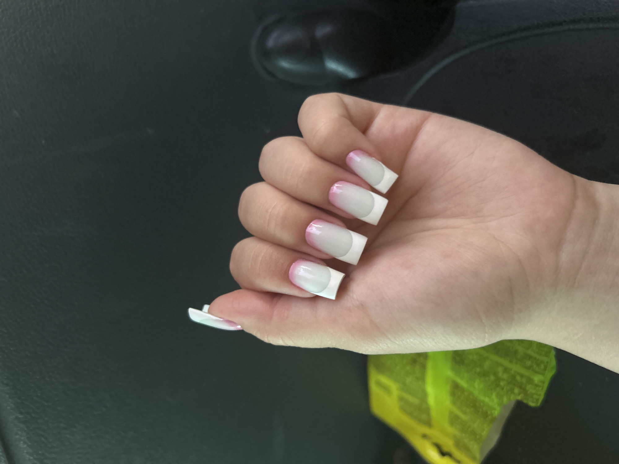 A close-up, high-angle shot shows a left hand with acrylic nails that have a French manicure design. The nails are square-shaped with white tips and a gradient of pink and white on the base. The hand is positioned in front of a dark, textured surface, possibly a car interior or furniture. To the left of the hand, a bright yellow object with a leaf-like pattern is partially visible. The background is dark and out of focus. There is no identifiable location in the city of Maiaky, Ukraine, as the image is cropped and lacks contextual details.