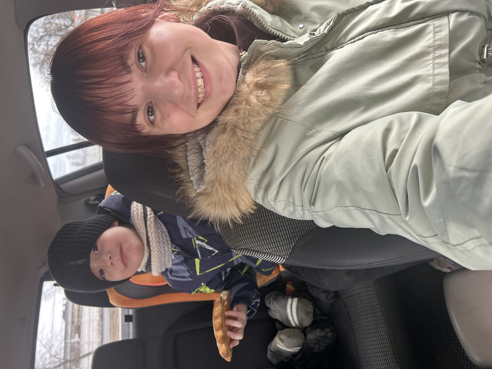 An adult woman and a young child are captured in a car selfie, likely taken in Chaikovsky, Russia. The woman, with reddish-brown hair and bangs, is smiling broadly at the camera while wearing a light green winter jacket featuring a prominent faux fur-lined hood. She is positioned in the front seat, tilting her head for the photo.
Behind her, a young child sits in a car seat with orange accents. The child wears a black knit beanie with "MONT" in white text, a dark blue winter jacket with a light-colored geometric pattern, and a light-colored knitted scarf. He also looks towards the camera with a slight smile and holds a long, golden-brown pastry or bread item in his right hand.
Through the car windows, a wintery landscape with bare trees and some snow is visible, indicating cold weather conditions. Grey mittens are seen on the floor or seat beside the child. The natural light filtering into the car suggests it is daytime. Both subjects appear to be in a pleasant mood.