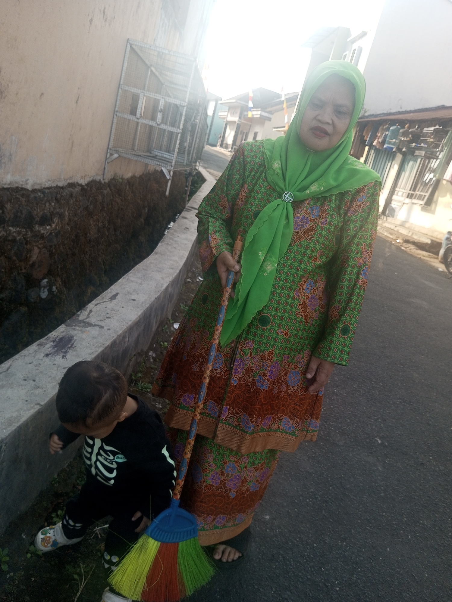 A woman wearing a green headscarf and a green and orange patterned dress stands on a paved road, holding a colorful broom. To her left, a young child in a black outfit with a skeleton print and white shoes looks down at the ground. A concrete wall with an attached metal cage structure is to the left of the woman and child. The scene appears to be in an outdoor urban environment in Krasak, Indonesia, with buildings visible in the background.