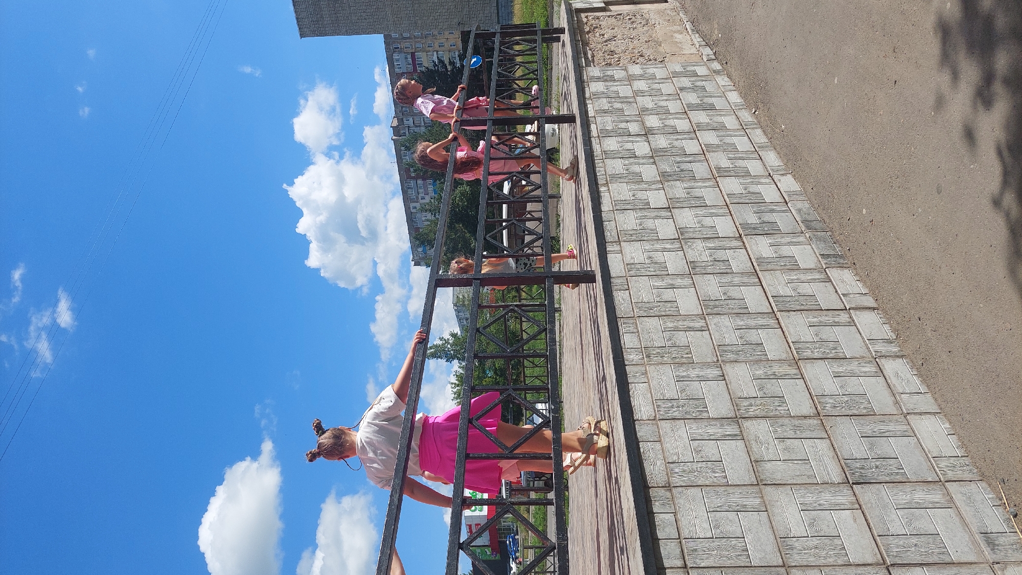 In Amursk, Russia, on a clear day with scattered clouds, a group of four young girls are observed on a paved outdoor area. The foreground features a wall constructed with rectangular grey tiles arranged in a patterned design. Behind this wall, a raised platform is visible, bordered by a black metal railing.
The girls, all wearing casual attire, are interacting with the railing. The girl closest to the viewer, positioned on the left side of the frame, has her back to the camera and is wearing a white t-shirt and a vibrant pink skirt. She is leaning against the railing with her right hand. To her right, a second girl in a pink dress is also leaning on the railing, facing away from the viewer. Further to the right, two more girls are present. One, wearing a pink dress, is looking towards the right of the frame, her body angled as if in motion. The fourth girl, on the far right, is also looking to the right, her posture suggesting movement.
In the background, a building with a grey facade stands, and beyond that, trees and more residential buildings can be seen. The sky above is a bright blue, indicative of fair weather. The presence of power lines overhead suggests an urban or suburban setting. The overall scene depicts a moment of outdoor activity for the children.