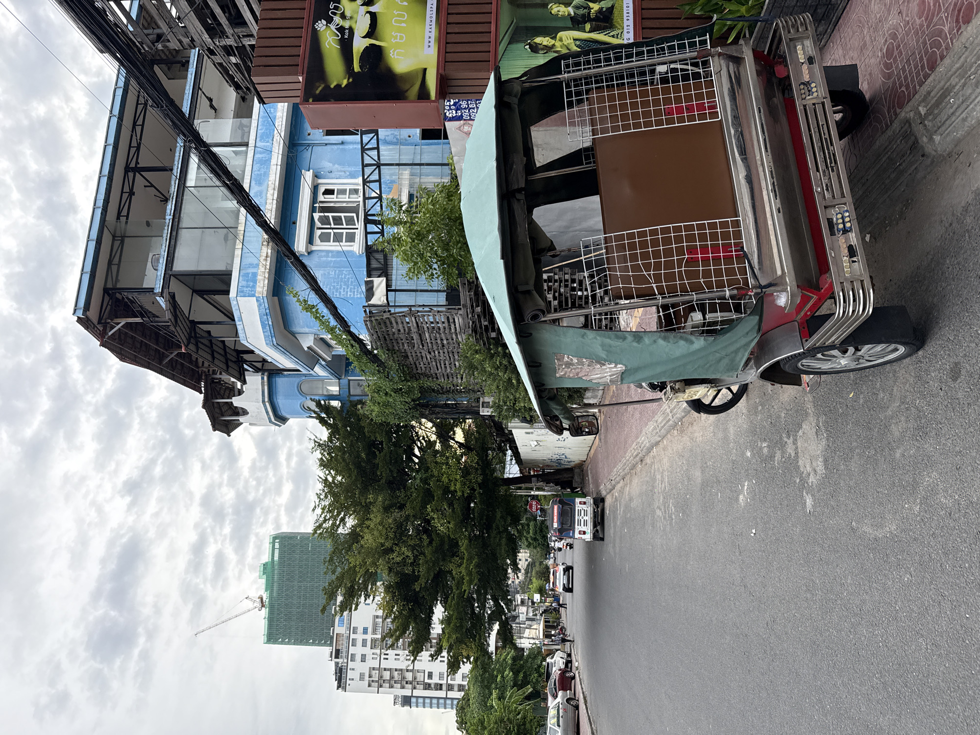 A street scene in Khan Daun Penh, Cambodia, features a tuk-tuk parked on the right side of the asphalt road, its green canopy and metal cage clearly visible. Adjacent to the road is a paved sidewalk with a red brick pattern, bordered by a low concrete curb. Beyond the sidewalk, a building with a bright blue facade stands out, featuring white-trimmed windows and multiple stories. The building's upper floors appear to be undergoing renovation or are designed with an open, modern aesthetic, with exposed metal beams and glass panels. In the background, a tall, modern building, partly covered in green scaffolding, dominates the skyline, with a construction crane extending from its top. Several other buildings and trees line the street, which shows moderate traffic, including cars and other tuk-tuks. The sky is overcast, with diffused light. A large advertisement featuring a woman and Cambodian script is mounted on a brown structure to the right of the blue building.