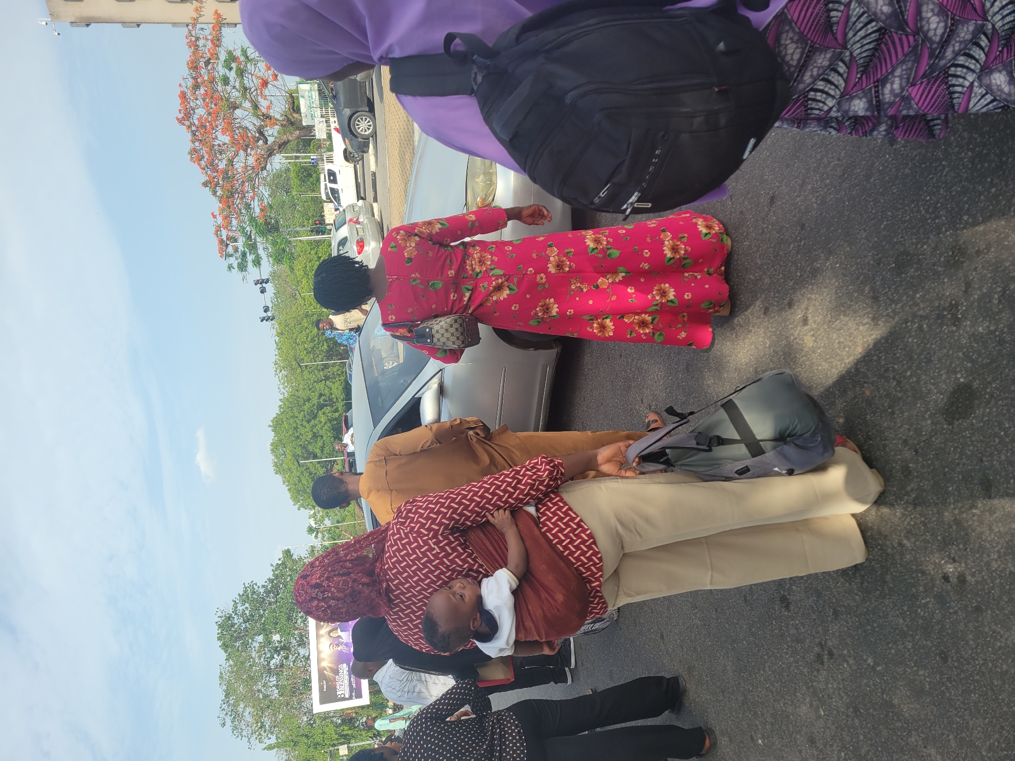 The image shows a group of people on what appears to be a road or street in Abuja, Nigeria. There's a woman in a red floral dress standing near a silver car. Another woman in a red patterned top is holding a child in a carrier. Nearby, a man is dressed in brown. A large advertisement board is visible in the background. The sky is blue, and there are trees in the vicinity.