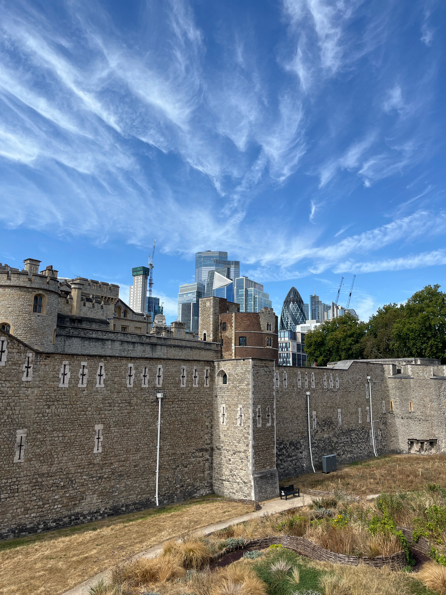 The image depicts a view of the Tower of London, a historic castle located on the north bank of the River Thames in central London, United Kingdom. The foreground is dominated by the ancient stone walls of the Tower, with small, cross-shaped window openings visible. The walls are made of weathered, light-colored stone, suggesting their age. To the right of the stone walls, there is a section of a more rounded, reddish-brown brick tower, and beyond that, more stone fortifications.
The ground in front of the walls is covered in dry, golden-brown grass and some scattered plants, indicating a parched or arid environment. A narrow, paved pathway winds through this area, and a dark park bench is visible near the wall.
In the background, a stark contrast is presented by a cluster of modern skyscrapers, defining the city's skyline. These buildings are of various heights and architectural styles, some with reflective glass facades and angular designs. Notable among them is the distinctive Gherkin building, with its unique rounded shape. Construction cranes are also visible among the taller buildings, suggesting ongoing development in the area.
Above the cityscape, a bright blue sky is filled with wispy, white cirrus clouds, creating a dynamic and textured canopy. The sunlight appears to be strong, casting shadows and illuminating the scene. The overall impression is a juxtaposition of historical architecture with contemporary urban development, set against a bright, cloudy sky.