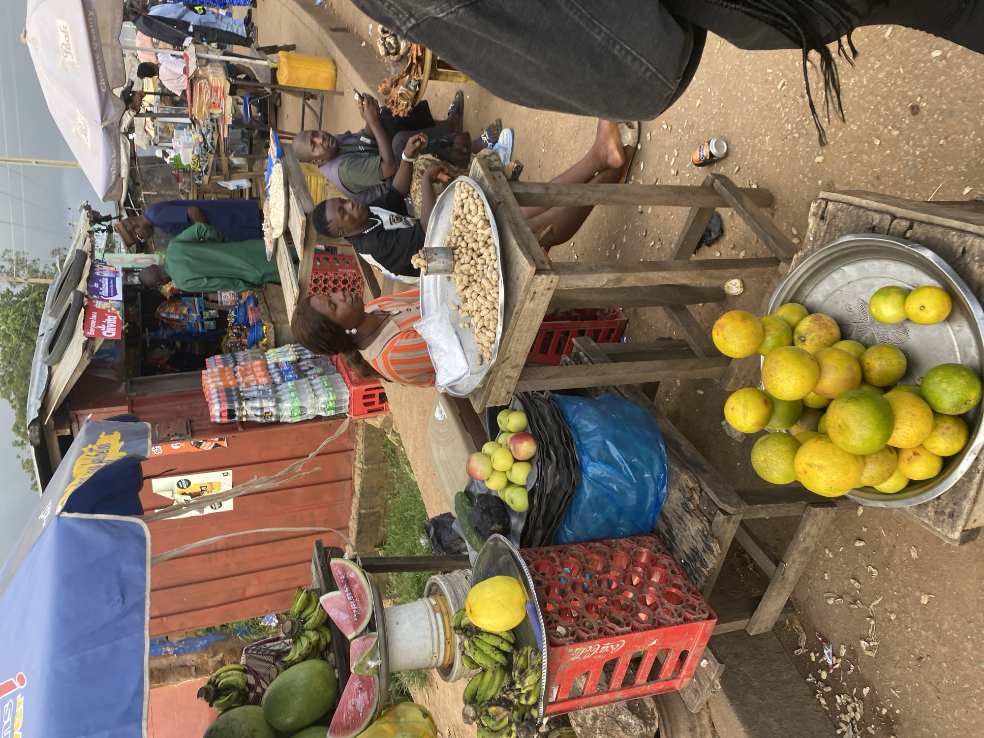 The image depicts a bustling outdoor market scene in Bukuru, Nigeria. In the foreground, a display of vibrant yellow and green citrus fruits is presented on a metal tray, some stacked in pyramids. To the left, a red Coca-Cola crate holds a bunch of bananas. Behind this, a pile of watermelons, some sliced, rests on a tiered stand. A woman in an orange and white striped shirt sits at a wooden table laden with a large tray of peanuts in their shells, accompanied by a metal cup. Next to her, two men are seated; one in a black and white t-shirt, the other in a green vest, is looking at his phone. In the background, various market stalls are visible, displaying goods like bottled drinks and snacks under umbrellas and makeshift roofing. People are seen browsing and interacting at the stalls. The ground is dusty and scattered with debris, indicative of a busy, informal marketplace. The overall atmosphere is one of lively commerce and everyday life in a Nigerian city.