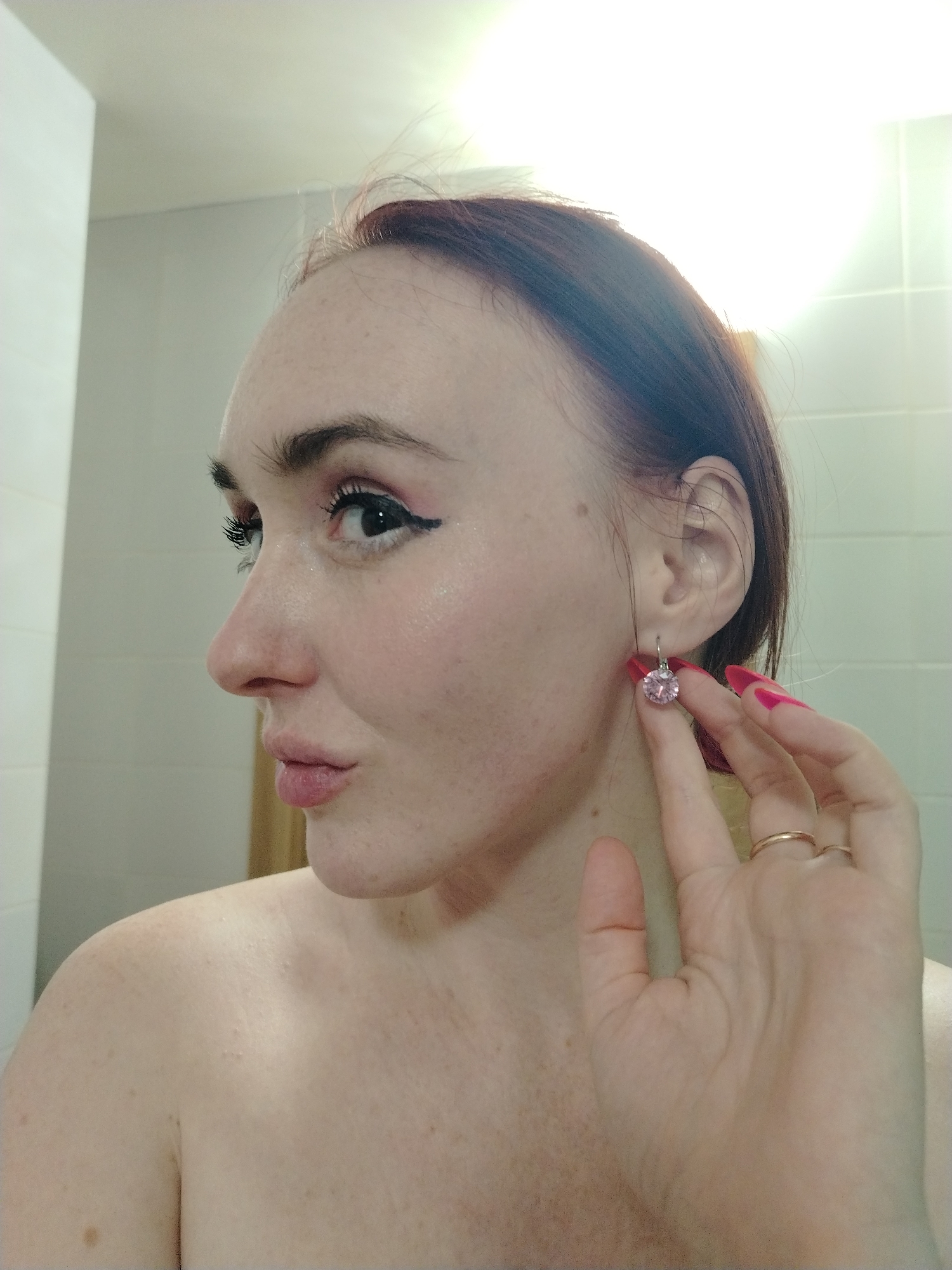 A fair-skinned woman with auburn hair is posing in a bathroom with white tile walls, likely in front of a mirror. She is seen from the shoulders up, facing left in profile with her head turned towards the camera. She is wearing a pink gemstone earring, which she is touching with the fingers of her right hand. She is wearing two gold rings on her right hand. She has dark eyeliner and pink lipstick. No evidence suggests this photo was taken in Izhevsk, Russia.