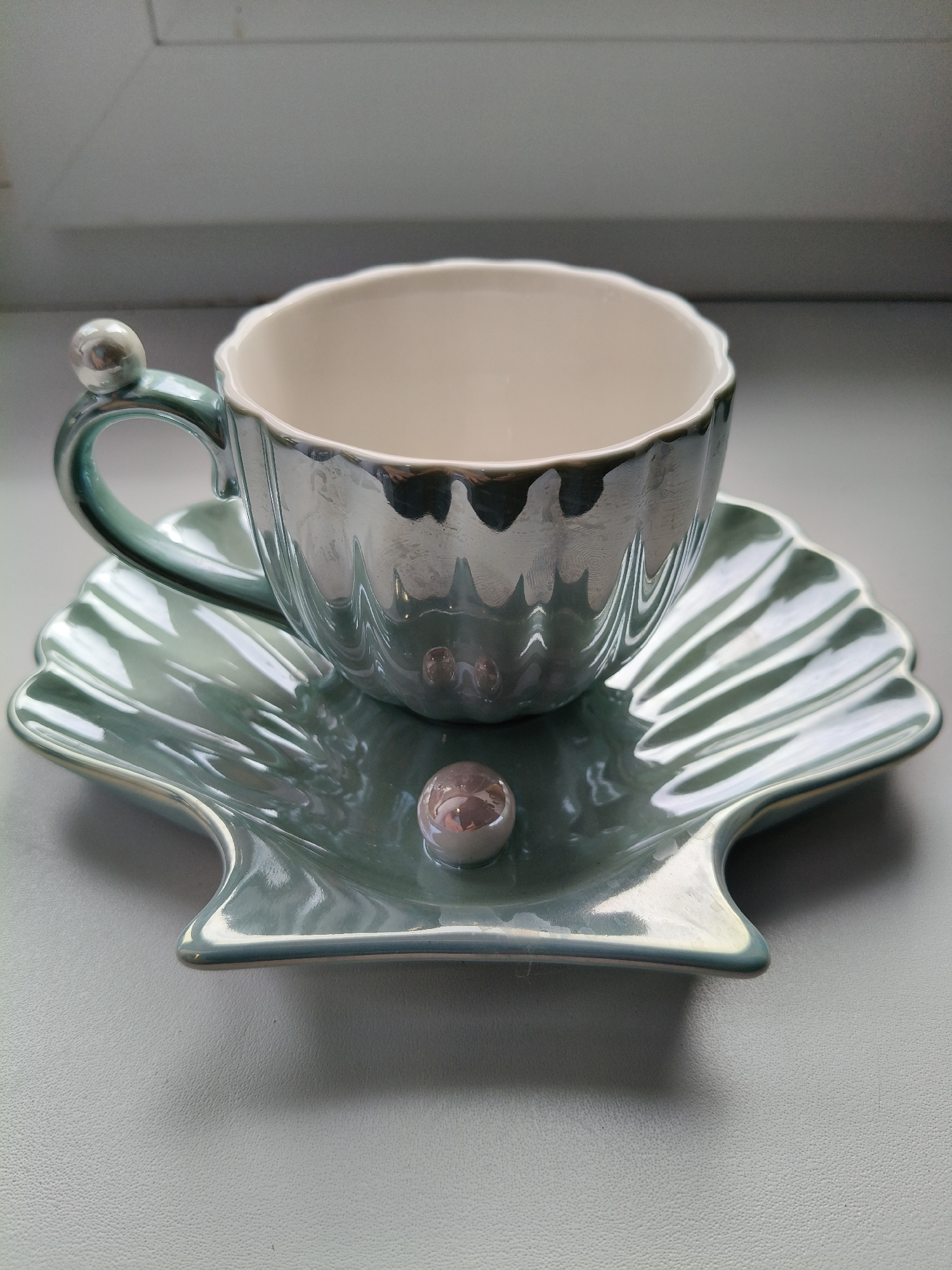 This is a close-up, eye-level, indoor photograph taken in Bigauņciems, Latvia. The image features a decorative teacup and saucer set, designed to resemble a seashell. The teacup has a fluted, iridescent exterior in shades of light blue and silver, with a cream-colored interior. Its handle is curved and adorned with a small, pearl-like embellishment at the top. The saucer is also shell-shaped, with raised ridges radiating from the center, and shares the same iridescent blue-green and silver finish as the cup. A small, translucent, pearl-like sphere rests on the saucer in front of the cup. The teacup and saucer are positioned on a light gray, textured surface, with a white surface visible in the background, suggesting it might be a windowsill or shelf. There are no people or animals present in the image.