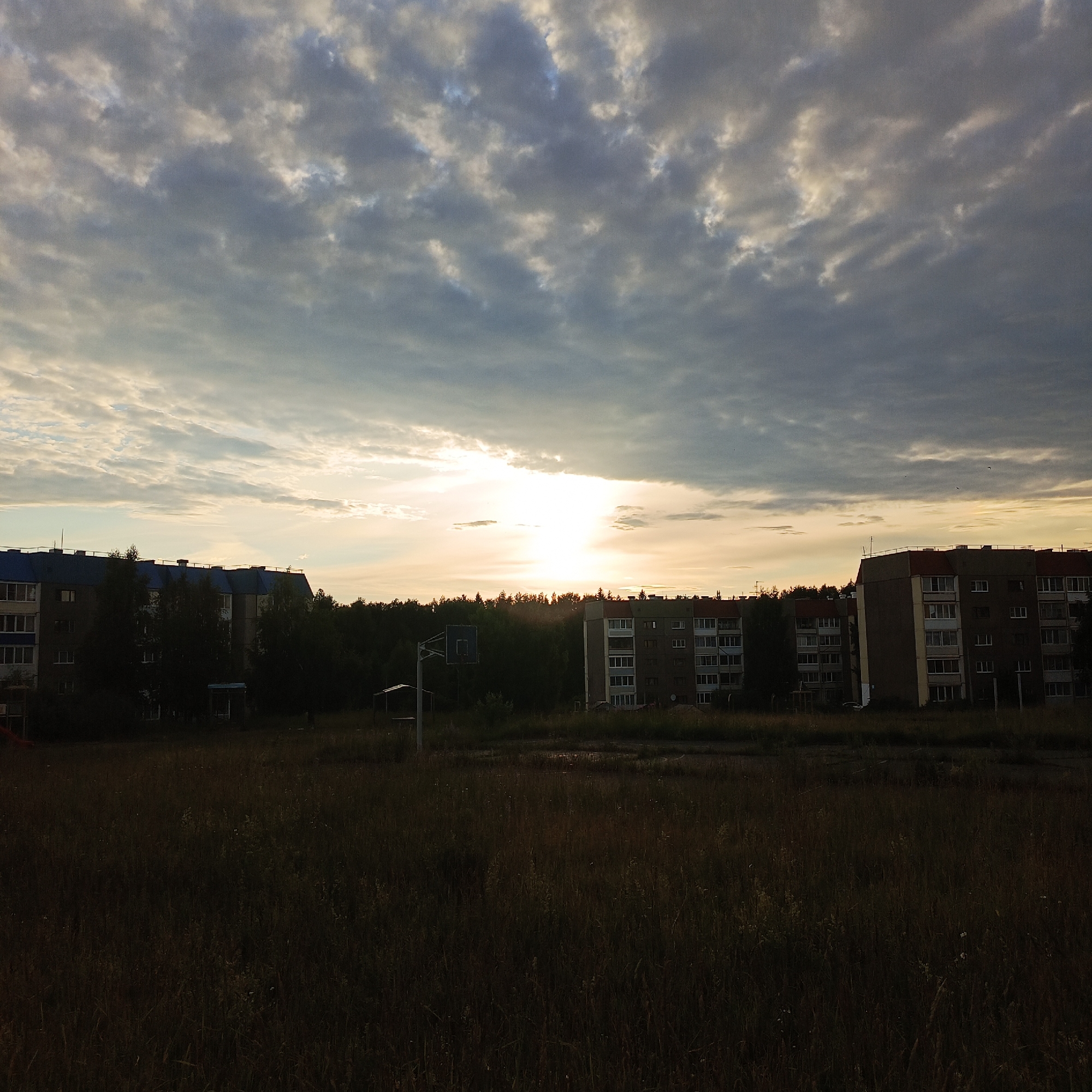 This is a landscape photograph captured at dusk in Марковский, Russia. The image depicts a residential area with several multi-story buildings silhouetted against a dramatic sky. The sun is setting, casting a warm, golden glow through a textured layer of clouds that dominate the upper half of the frame. The clouds range in color from dark gray to bright white, indicating a mix of light and shadow.
In the foreground, a field of tall, dry grass recedes towards the buildings, creating a sense of depth. The grass is dark and somewhat out of focus, emphasizing the light in the sky and the buildings. Among the grass, a basketball hoop with a backboard is visible, along with some small, covered structures, suggesting a recreational area.
The buildings themselves are typical of Soviet-era apartment blocks, with flat roofs and numerous windows. Some of the buildings have dark blue or red roofing, providing a contrast to the muted tones of the rest of the scene. The trees in front of the buildings are dark and dense, appearing as a solid line of foliage.
The overall mood of the image is peaceful and serene, with a touch of melancholy due to the fading light. The time of day is clearly sunset or twilight, given the low angle of the sun and the warm, diffused light. The weather appears to be partly cloudy, contributing to the striking visual of the sky. No text is visible in the image.