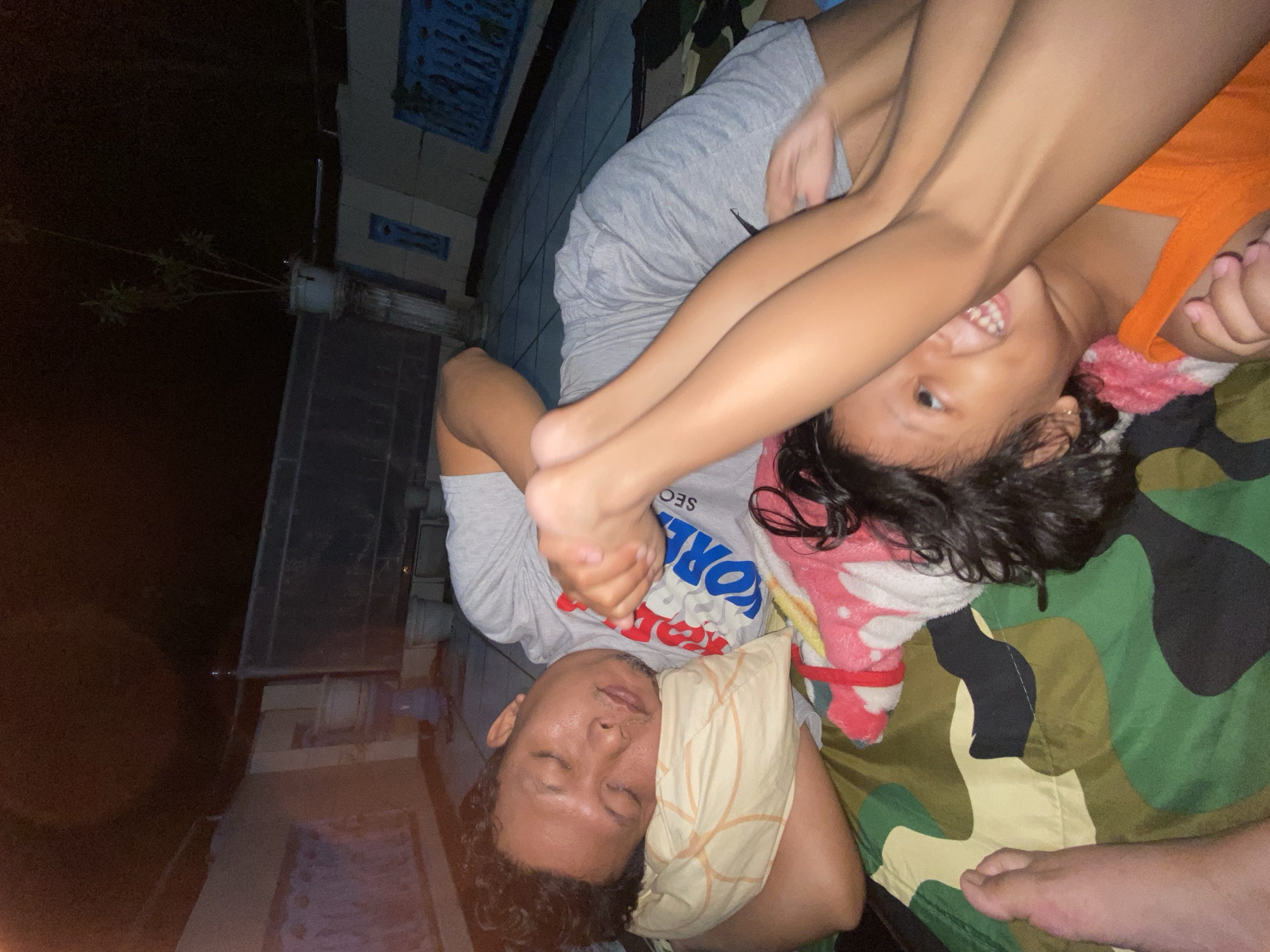 The media file shows a close-up, rotated view of two people lying down on a camouflage-patterned blanket, likely outdoors at night in Gelam, Indonesia. The person in the foreground is a man with his eyes closed, resting his head on a pillow. He has curly dark hair and stubble. He is wearing a grey t-shirt with text on it. To his right, a child is lying on top of him, with their legs in the air and their head resting on the man's chest area. The child is wearing an orange top. Their facial expression suggests they are smiling and looking towards the camera. There are hints of a tiled wall and some greenery in the background, suggesting a slightly unkempt outdoor area. The lighting indicates it is nighttime.