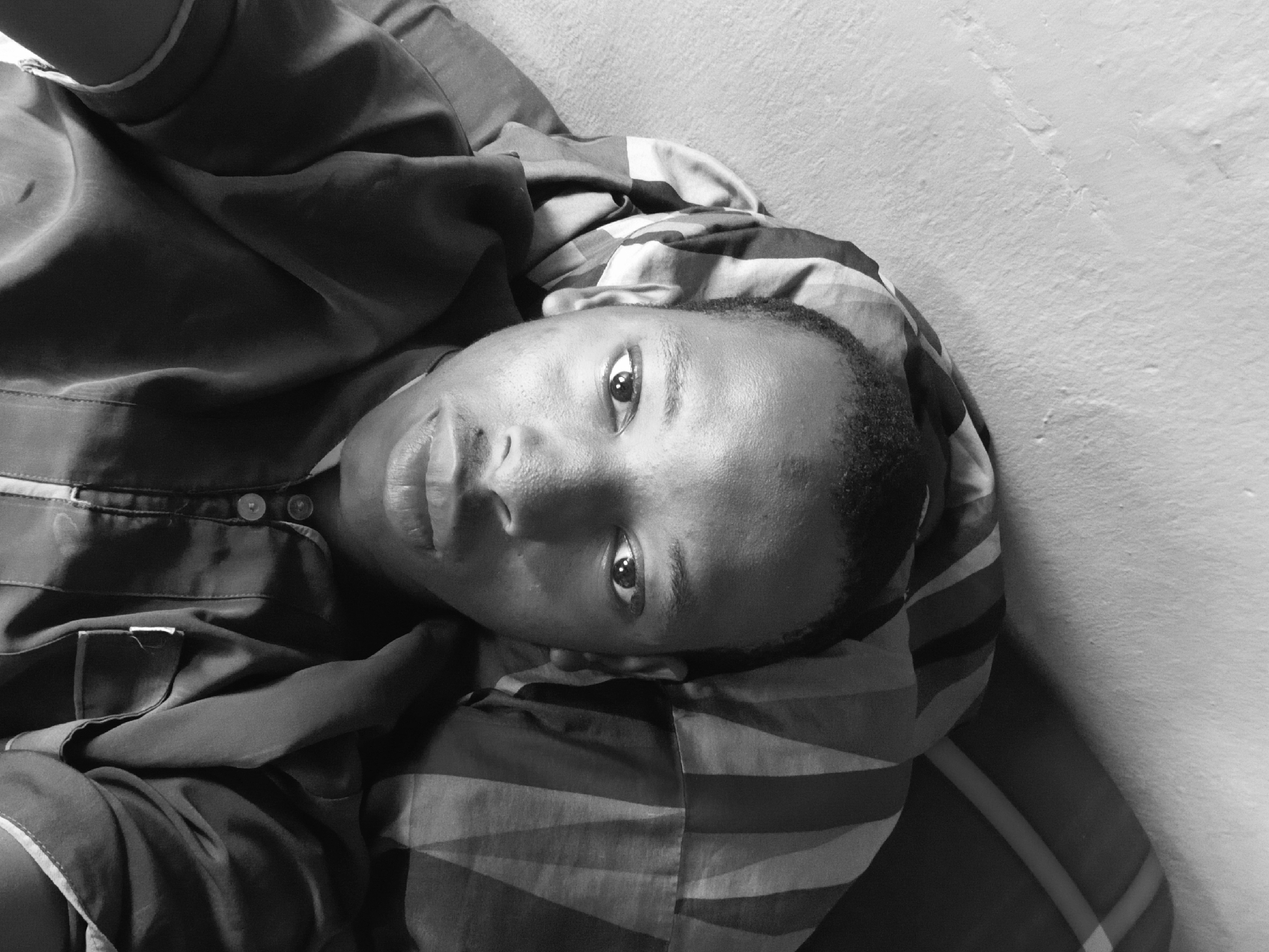 A person lies down, face towards the camera. The person is wearing a buttoned shirt and is lying on what appears to be a striped pillow or blanket. The background consists of a plain, light-colored wall. The image is in black and white. The photograph was taken indoors and there is not enough information to determine if the location is Lagos, Nigeria.