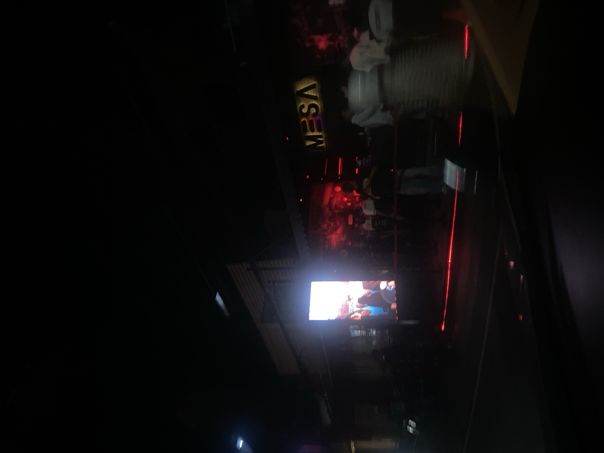 The image depicts a nighttime scene outside a bar or restaurant named "Mesa" in North Kuta, Indonesia. The establishment has a brightly lit sign. There's an outdoor area with people present; some are seated, others are standing. A large screen displays content, though the specific content is indistinct. Red lighting is visible within the venue. The photo was likely taken from inside a vehicle.