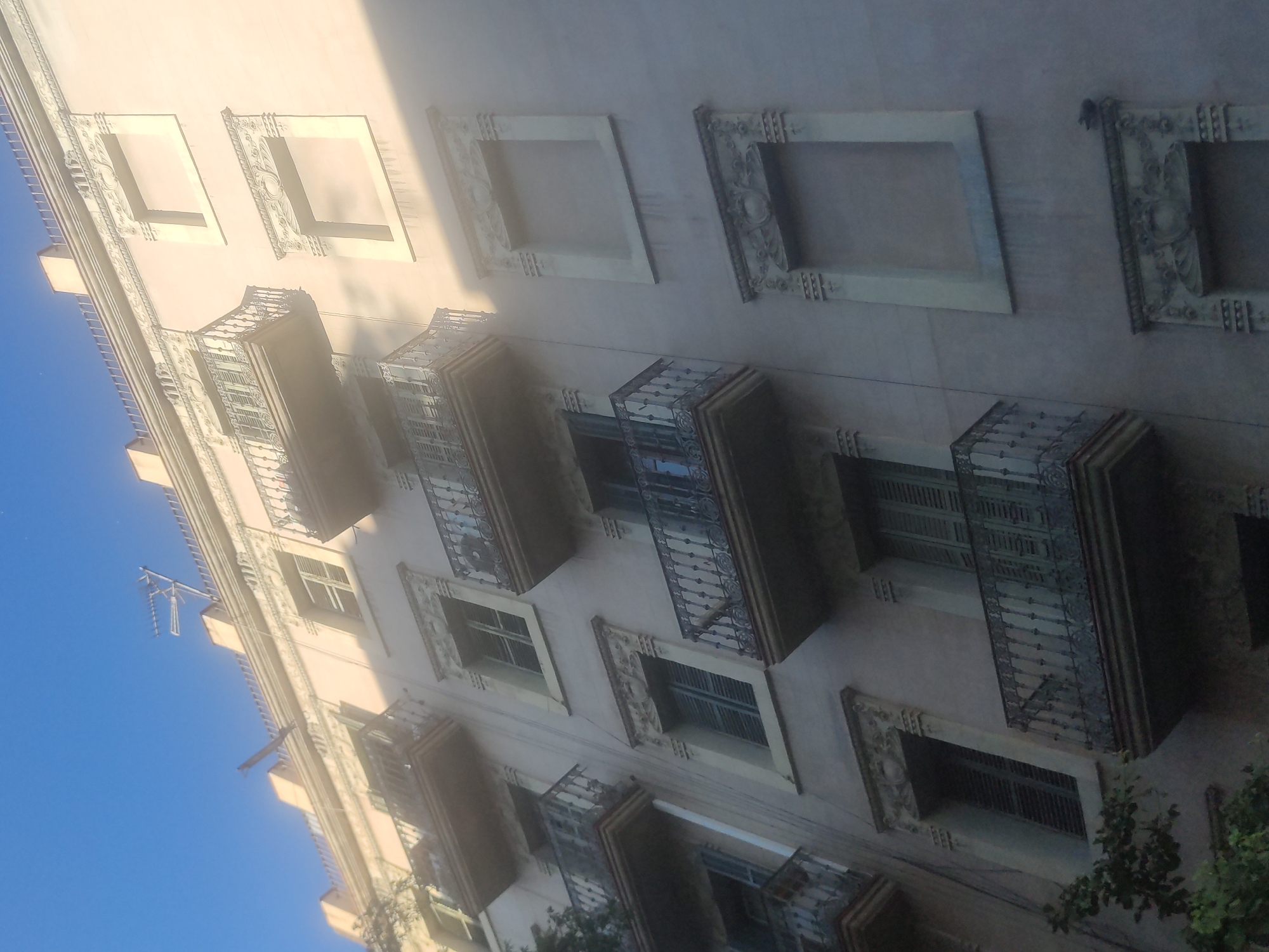 The image shows the facade of a building. The building has a light pink exterior with decorative window frames and wrought-iron balconies. The balconies are rectangular and project outward from the building. The windows have louvered shutters that are partially closed. The building is partially lit by direct sunlight, which casts shadows across the facade. The sky is visible in the lower left corner, and it is a clear blue. A small portion of foliage is visible in the lower right corner. An antenna is visible on the building.
