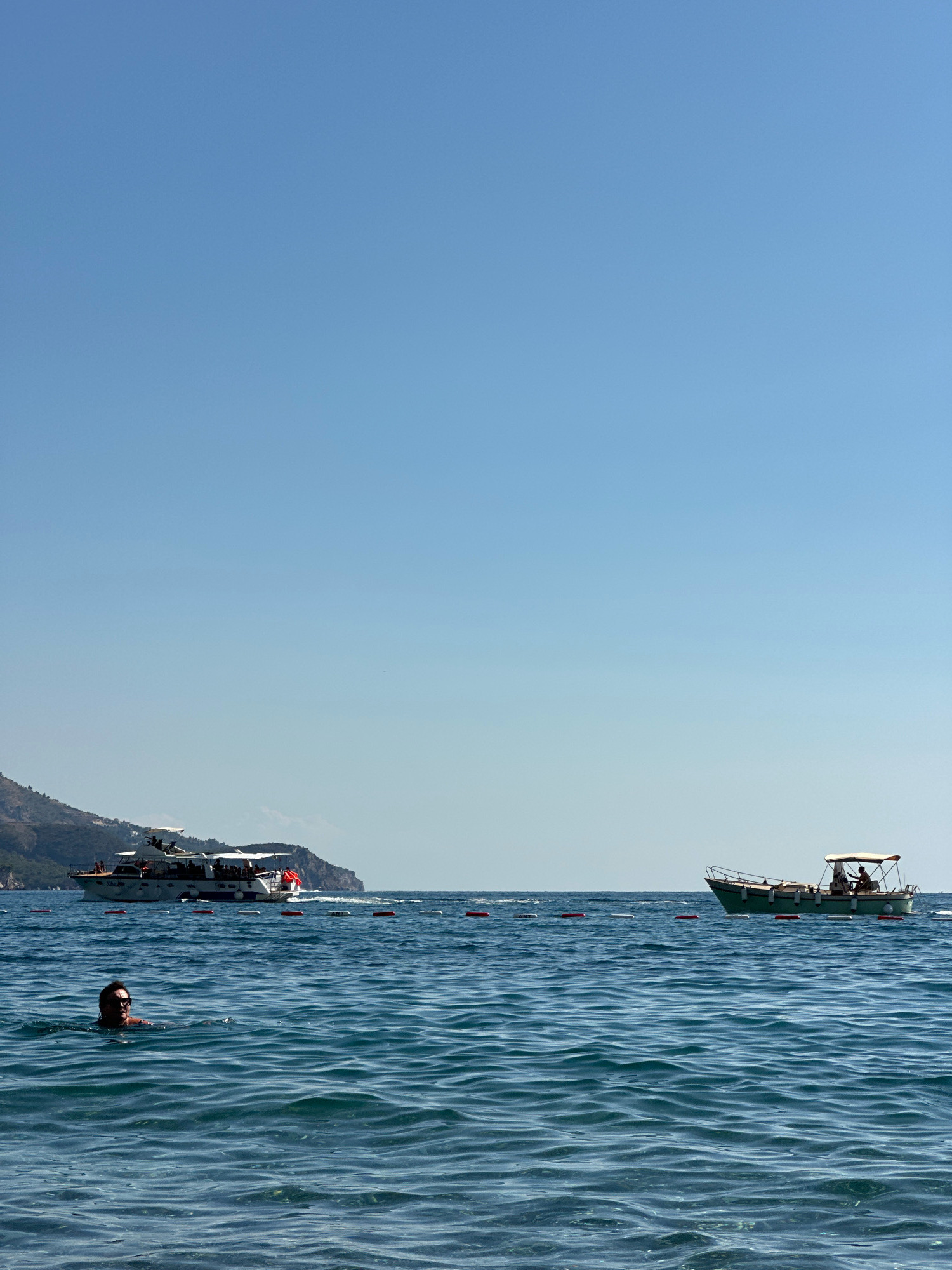 In the clear blue waters off the coast of Boreti, Montenegro, a lone swimmer surfaces, their head and shoulders visible against the rippling sea. In the distance, two boats navigate the calm waters. The larger vessel, a passenger boat, is closer to the shore, its white hull contrasting with the darker, mountainous landscape behind it. A smaller, green boat is further out, its canopy casting a shadow over its occupants. A line of buoys marks a boundary in the water, separating different areas of the sea. The sky above is a vast expanse of clear, unblemished blue, offering a serene backdrop to the maritime scene.