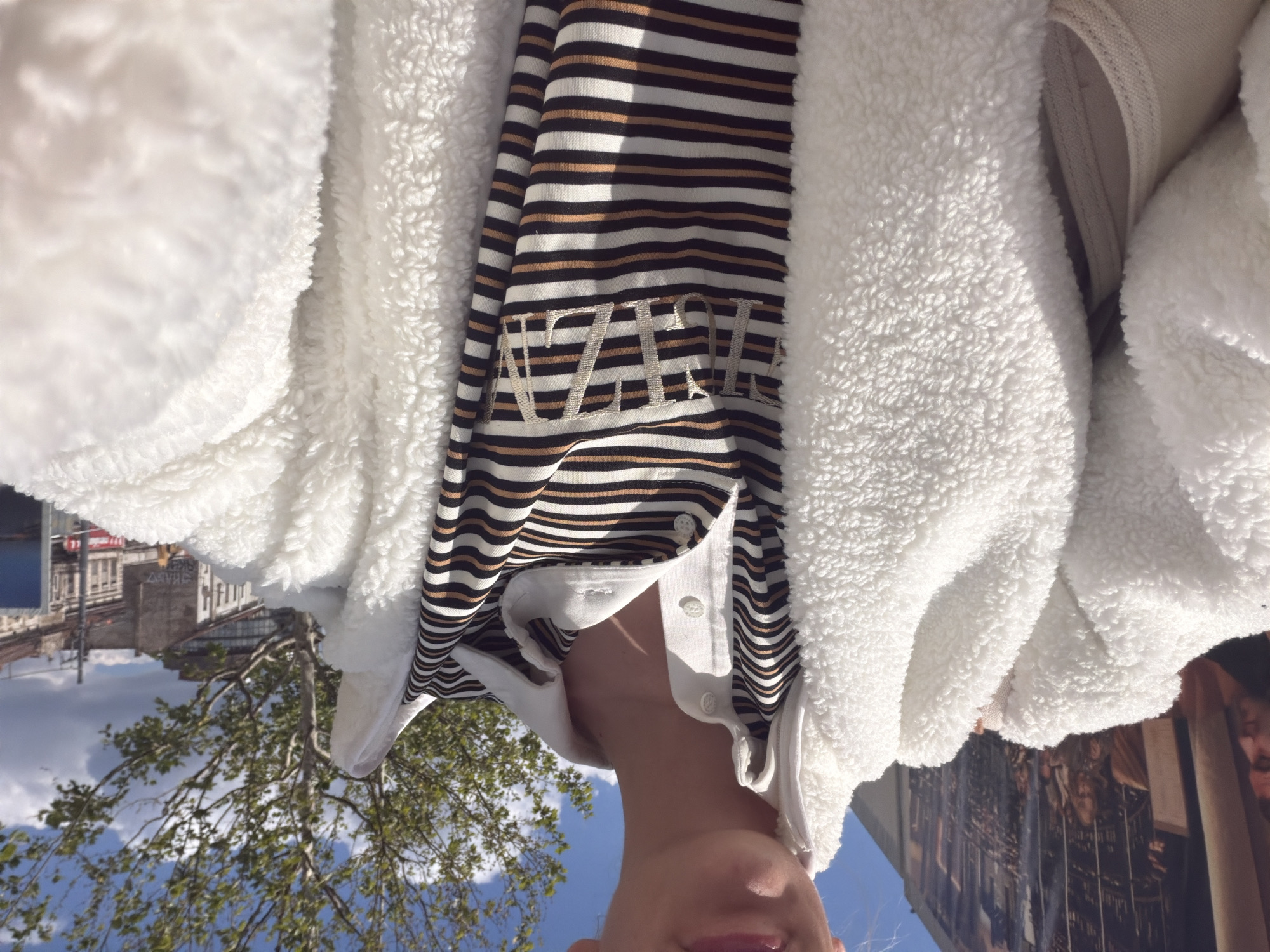 A person wearing a striped shirt and a white fleece jacket stands outside on a sunny day. In the background, trees and buildings are visible. "AZIZOL" is embroidered on the shirt. Buildings with advertisements and architecture typical of Belgrade are seen. A sign with Cyrillic writing suggests the scene is in Serbia.