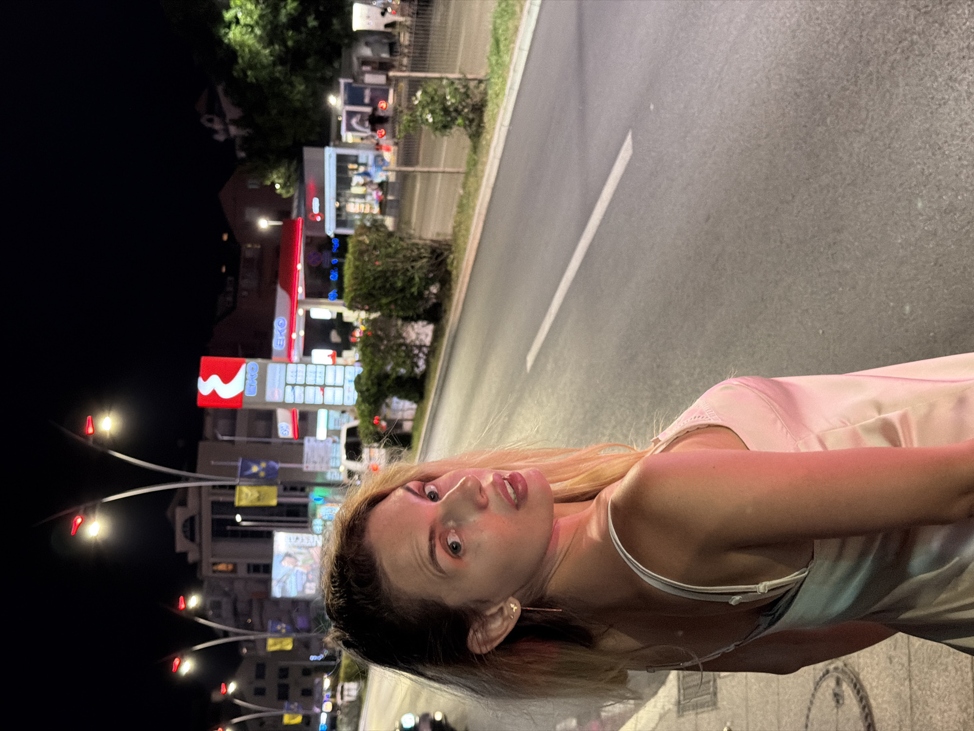 A young woman with blonde hair is walking at night along a street in Budva, Montenegro. She is wearing a light pink slip dress and looking over her left shoulder at the camera. The street is paved with asphalt and has a white line marking the lane. In the background, there is a gas station with illuminated signs, trees, and buildings. The scene is lit by streetlights and the lights from the businesses.
