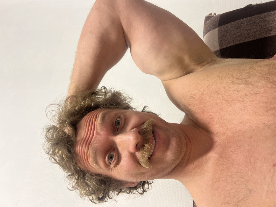 A man is depicted in a close-up, low-angle portrait, with the image rotated 90 degrees counter-clockwise. The man is looking directly at the camera with a friendly, broad smile, revealing his teeth. He has curly, light brownish-blonde hair that falls around his forehead and ears. A prominent, thick blonde or light brown mustache covers his upper lip. His eyes are light-colored, possibly blue or green. Numerous horizontal wrinkles are visible across his forehead, suggesting he might be raising his eyebrows.
He is shirtless, showcasing his bare chest and shoulders, which are covered in light body hair. His left arm is bent at the elbow and raised, with his hand seemingly placed on or behind his head, accentuating the musculature of his bicep. The pose and angle give the impression that he is either taking a selfie from a very low perspective or someone else is photographing him from below.
The background is predominantly a plain, light-colored surface, likely a wall or ceiling. In the top right corner of the original image (which would be the top right if viewed horizontally, or bottom right if rotated upright), a small section of a brown and dark-striped fabric object, possibly a blanket, pillow, or headboard, is visible. The overall mood of the image is lighthearted and cheerful due to his smiling expression. There is no visible text in the image.