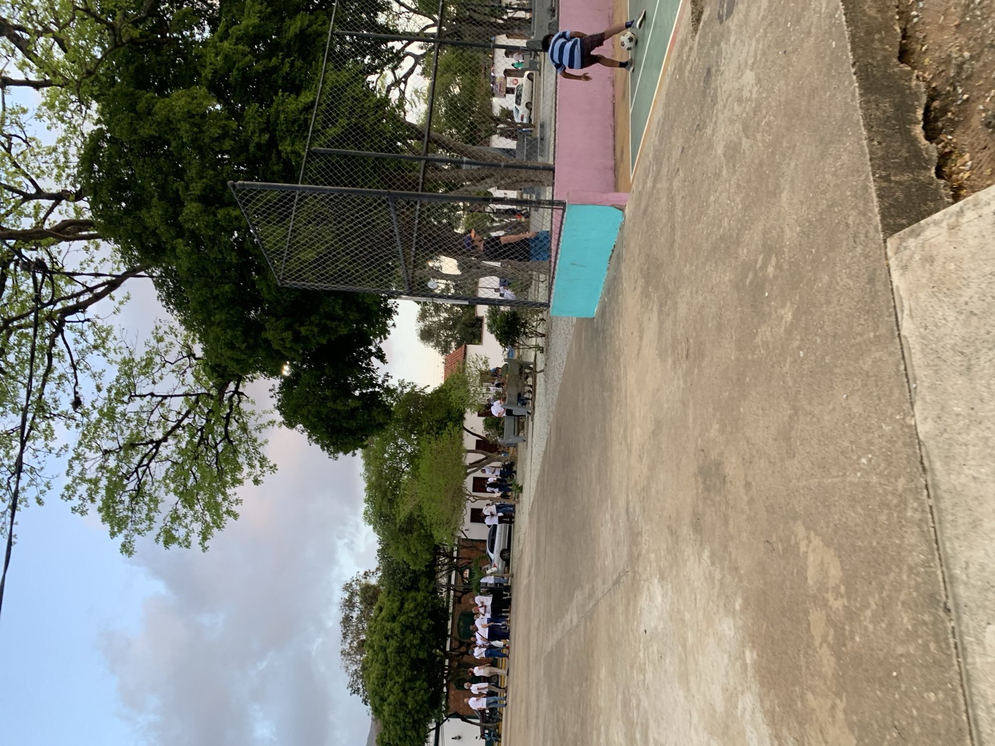 The image depicts an outdoor scene in a public space, possibly a park or plaza, in Santa Ana, Venezuela. In the foreground, a boy wearing a striped shirt and shorts is playing soccer on a court with a green surface bordered by pink and turquoise walls. A black metal fence encloses a portion of the area. In the background, a group of people, mostly wearing white shirts, are gathered near buildings with white walls and red-tiled roofs. Several trees with green foliage are present, and a silver car is parked nearby. The sky above is overcast with some clouds, suggesting it may be late afternoon or early evening. The ground in the foreground is made of concrete.