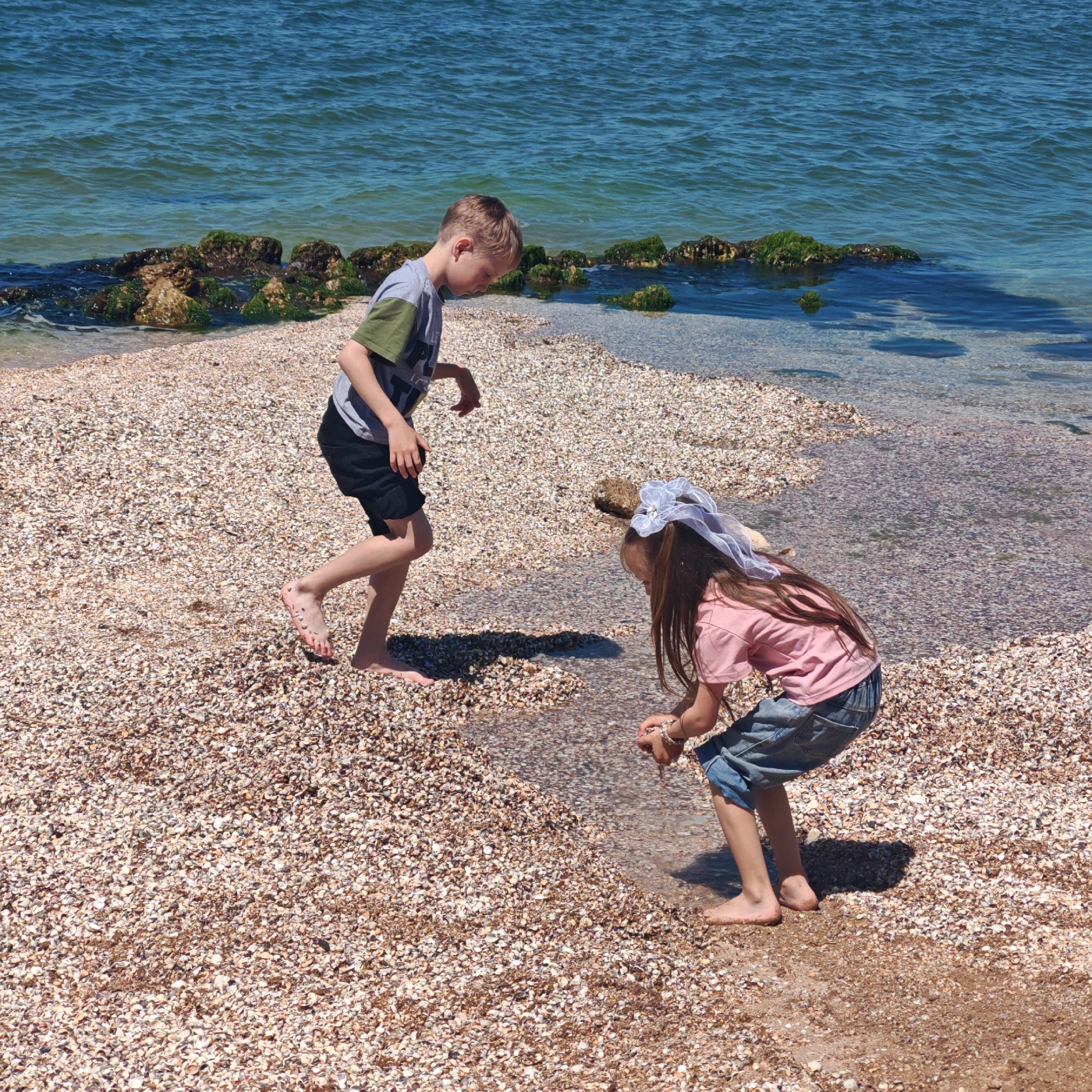 The image depicts two children on a beach in городское поселение Щелкино, Ukraine. A boy in a gray and olive green t-shirt and black shorts is walking barefoot on a beach covered in shells. A girl in a pink t-shirt and rolled-up denim jeans is bending down at the water's edge, possibly playing with something in her hands. She is wearing a white hair accessory. The sea is visible in the background, with rocks covered in green algae separating the beach.