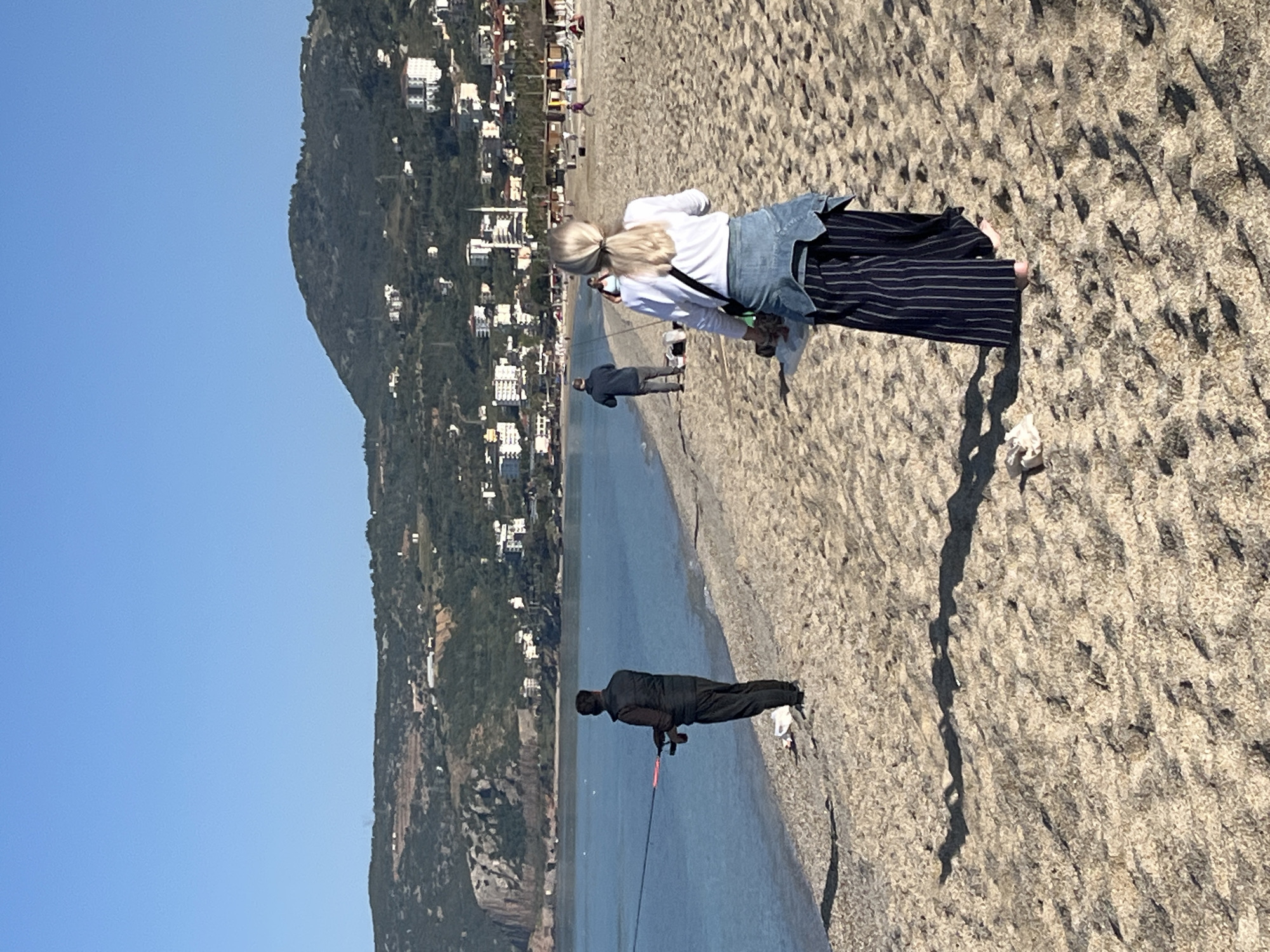 The image captures a beach scene in Alanya, Turkey, with a mountain providing a backdrop to the city's buildings. The sky is clear and blue. On the sandy beach, there are three individuals: a woman with blonde hair, wearing a white shirt, denim jacket, and striped pants, appears to be bending over. Further down the beach, a man in dark clothing is visible, holding a fishing rod. Another man in gray clothing is standing behind the woman. There is trash on the sand next to the woman. The water is calm, and the scene seems to capture a moment of leisure and fishing on the beach.