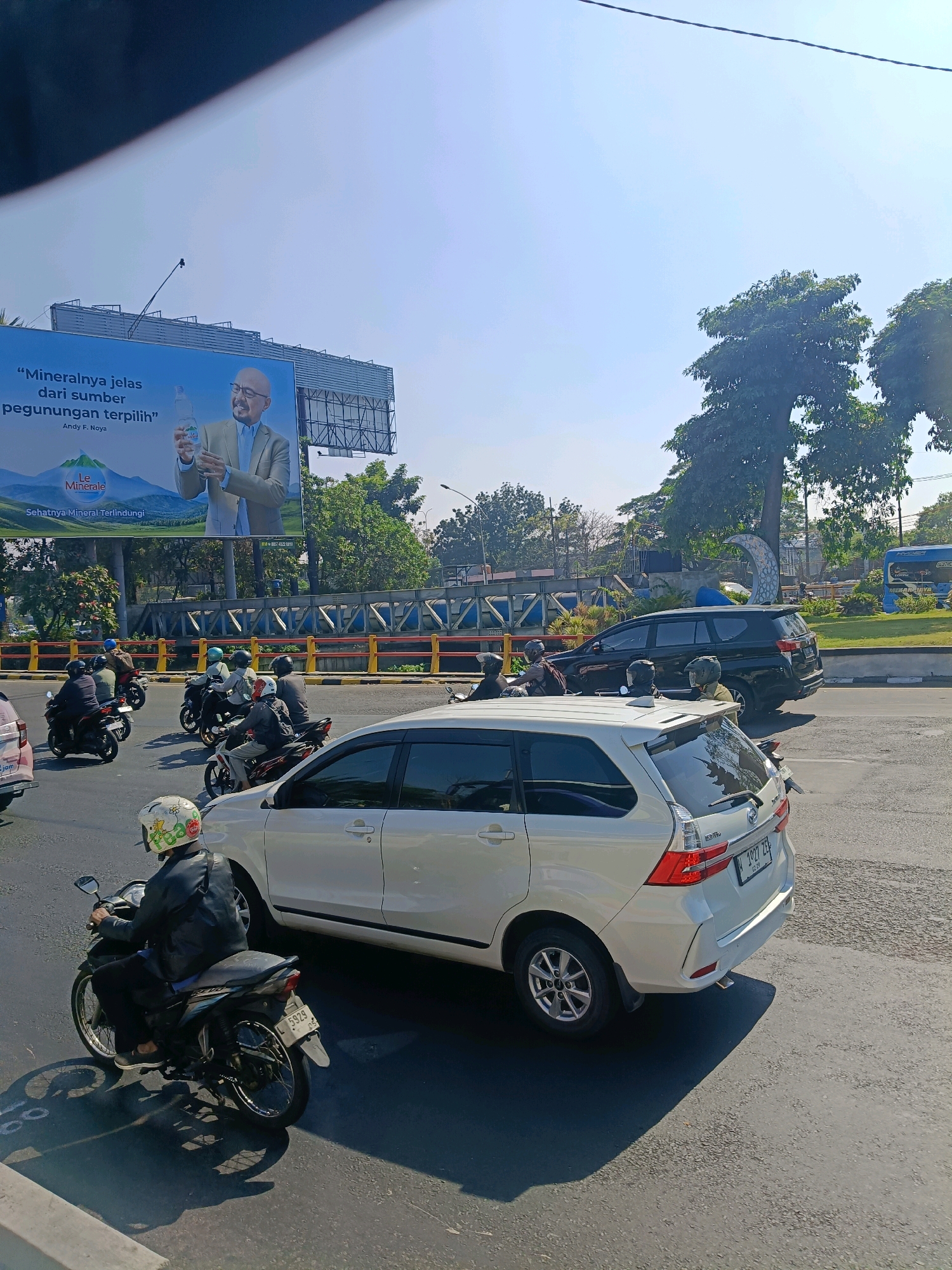 The image captures a street scene in Surabaya, Indonesia, on a clear, sunny day. The foreground is dominated by a white car, an MPV, in motion. To the left of the white car, a person on a motorcycle is visible, wearing a black jacket and a white helmet adorned with drawings. Behind them, several other motorcycles are also in traffic. In the background, a large billboard advertising "Le Minerale" mineral water is prominently displayed, featuring a man holding a bottle. Beyond the billboard, trees and street infrastructure, including a blue bus, are visible. The traffic appears to be moderate, with both cars and motorcycles navigating the road. The overall impression is of a typical urban environment with active street life under a bright sky.
